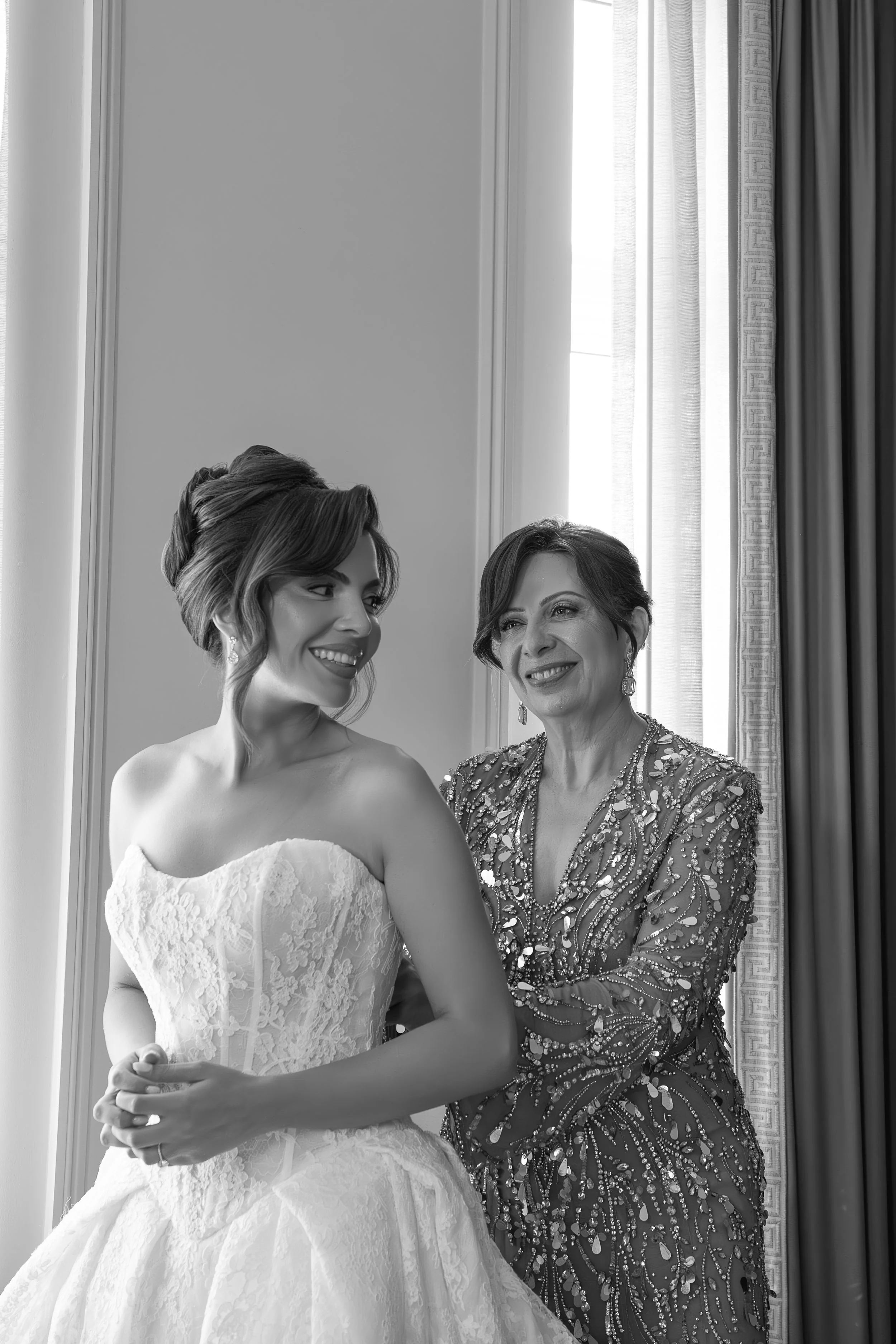A bride in a lace wedding gown standing with an older woman in a sparkling, beaded dress, both smiling, inside a room with curtains and a window.