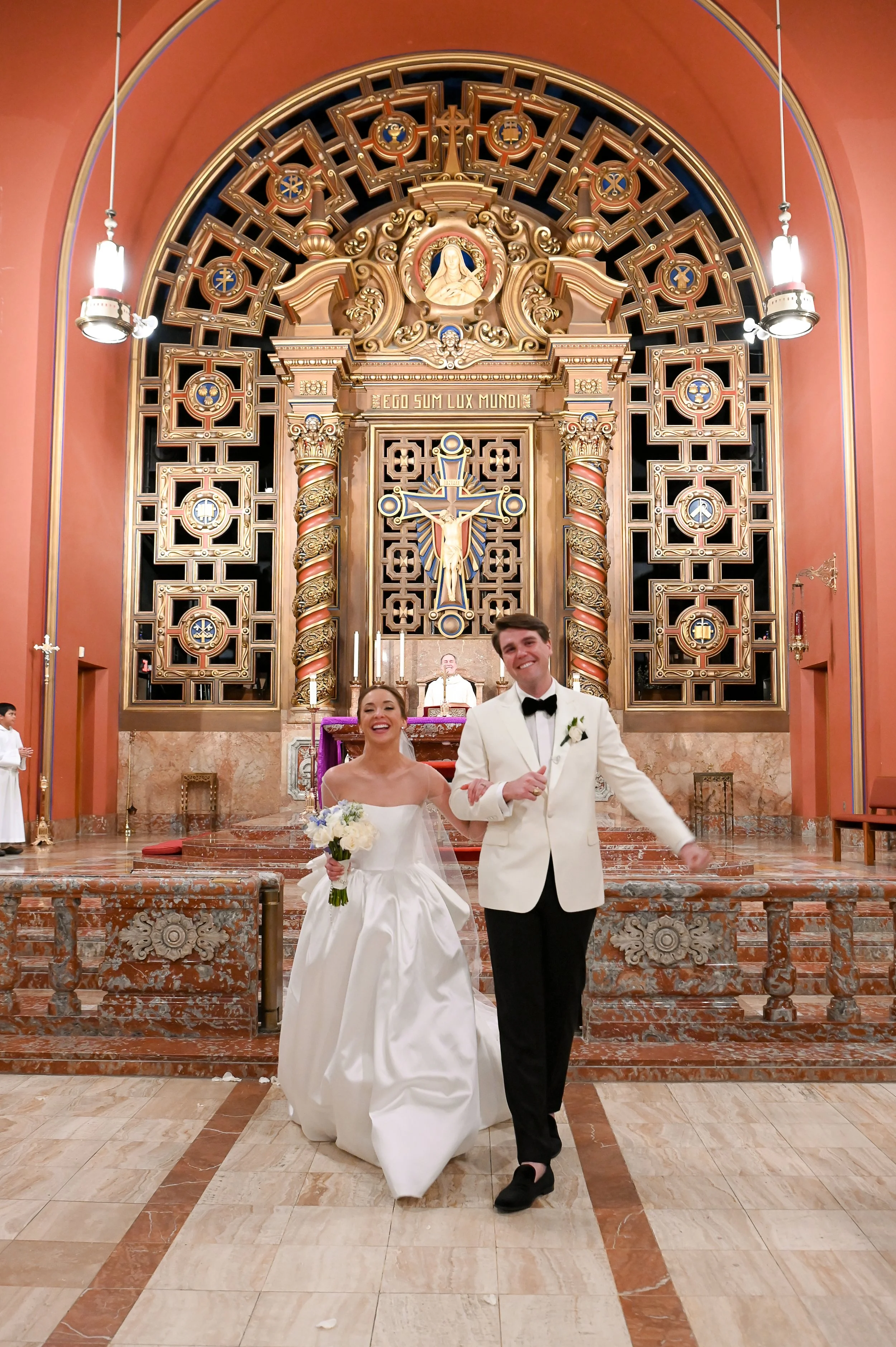 A newly married couple walking down the aisle of a church after their wedding ceremony. The bride is in a white wedding gown holding a bouquet, and the groom is in a white tuxedo jacket with a black bow tie. The church has ornate gold and wooden reli
