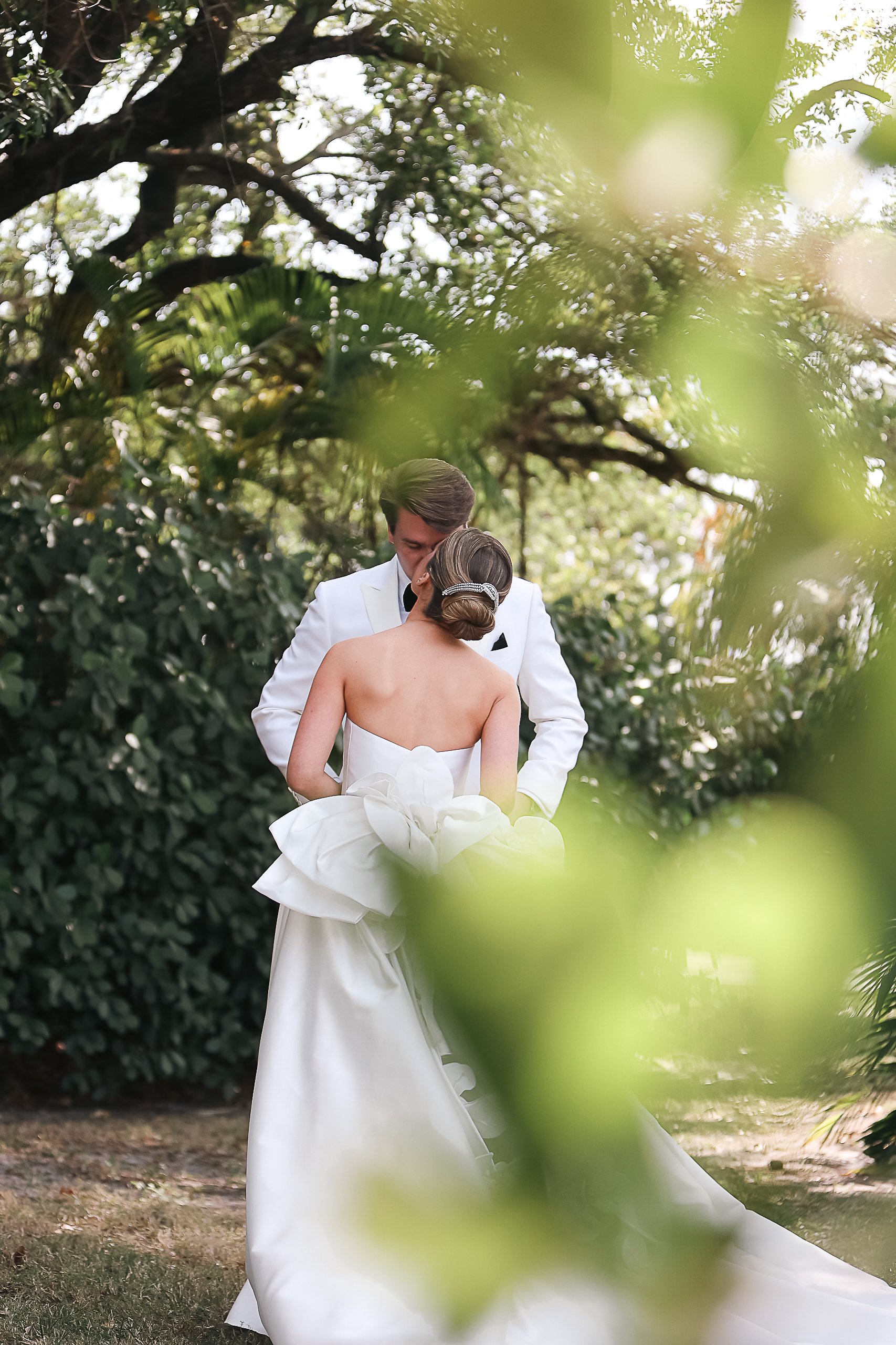 A bride and groom sharing a kiss outdoors, surrounded by trees and greenery, with a focus on the bride's back and wedding gown.
