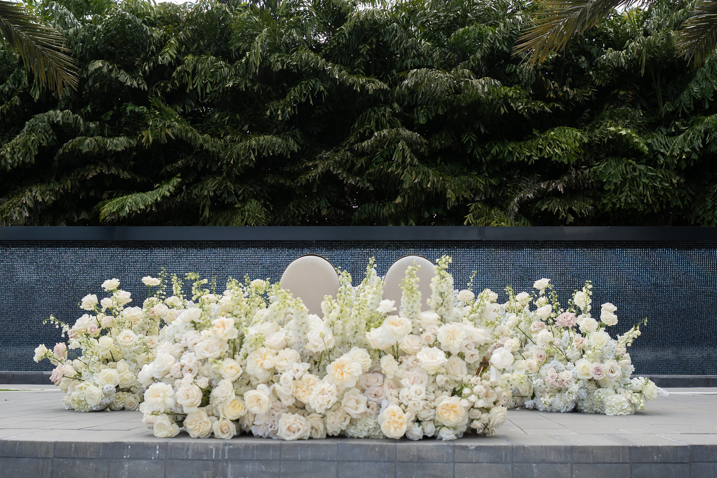 A large arrangement of white and pale pink flowers, possibly for a wedding, with a backdrop of green tropical leaves and a dark textured wall.