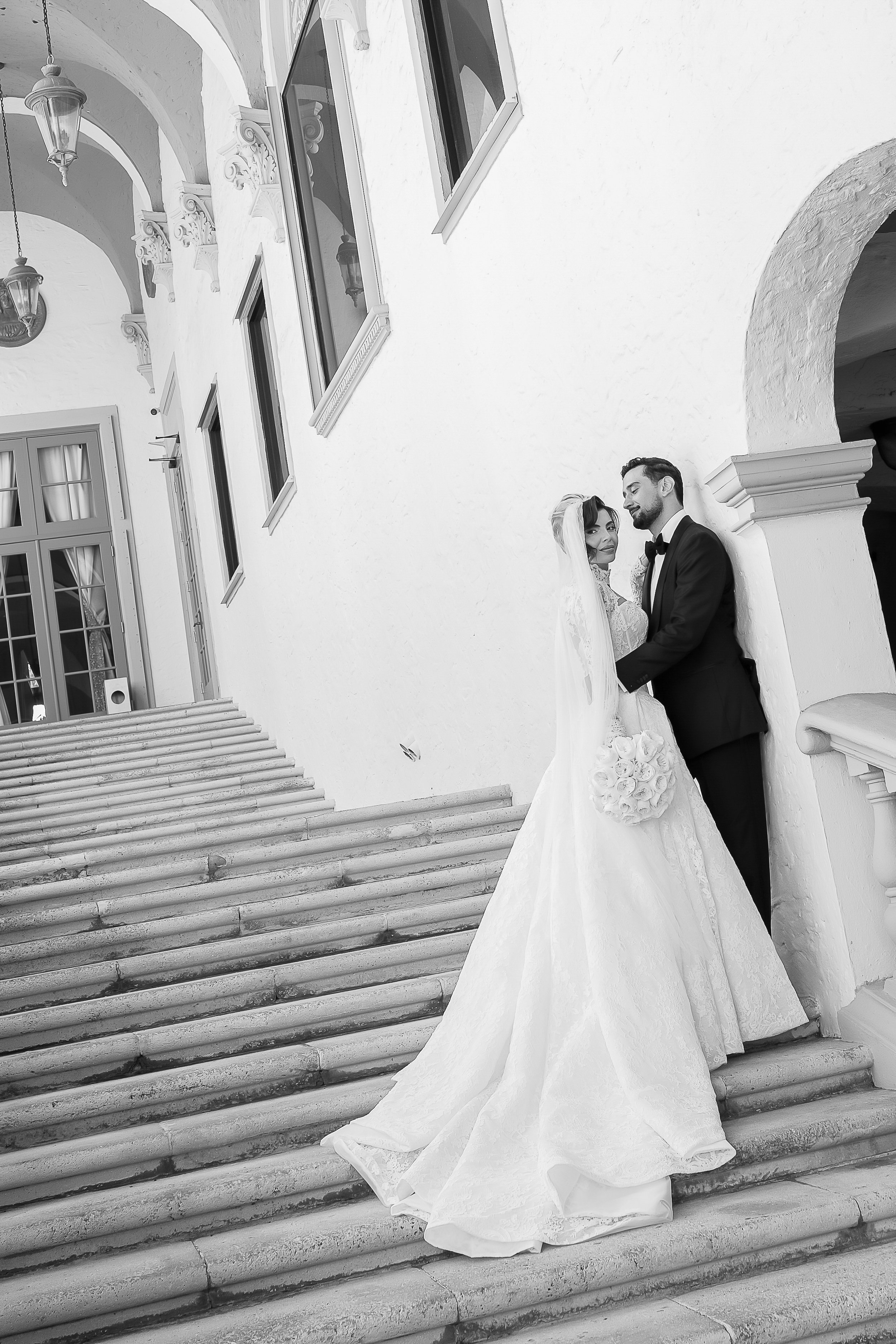 Black and white photo of a bride and groom standing on outdoor stairs against a white wall with windows. The bride is wearing a long wedding dress with train and holding a bouquet. The groom is in a tuxedo with bow tie, holding the bride's hand and l