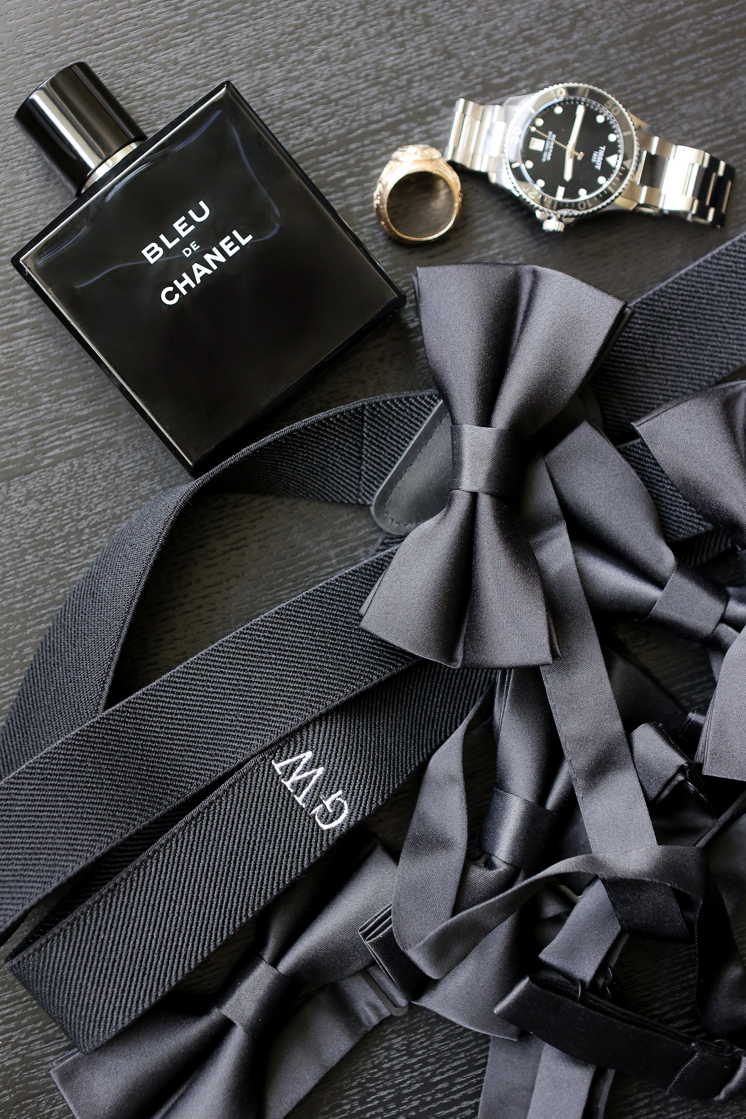 A black and gray tuxedo belt with a large bow, a bottle of Bleu de Chanel cologne, a wristwatch with a metal band, a gold ring, and a black box with the number 145 embroidered in white, on a dark surface.