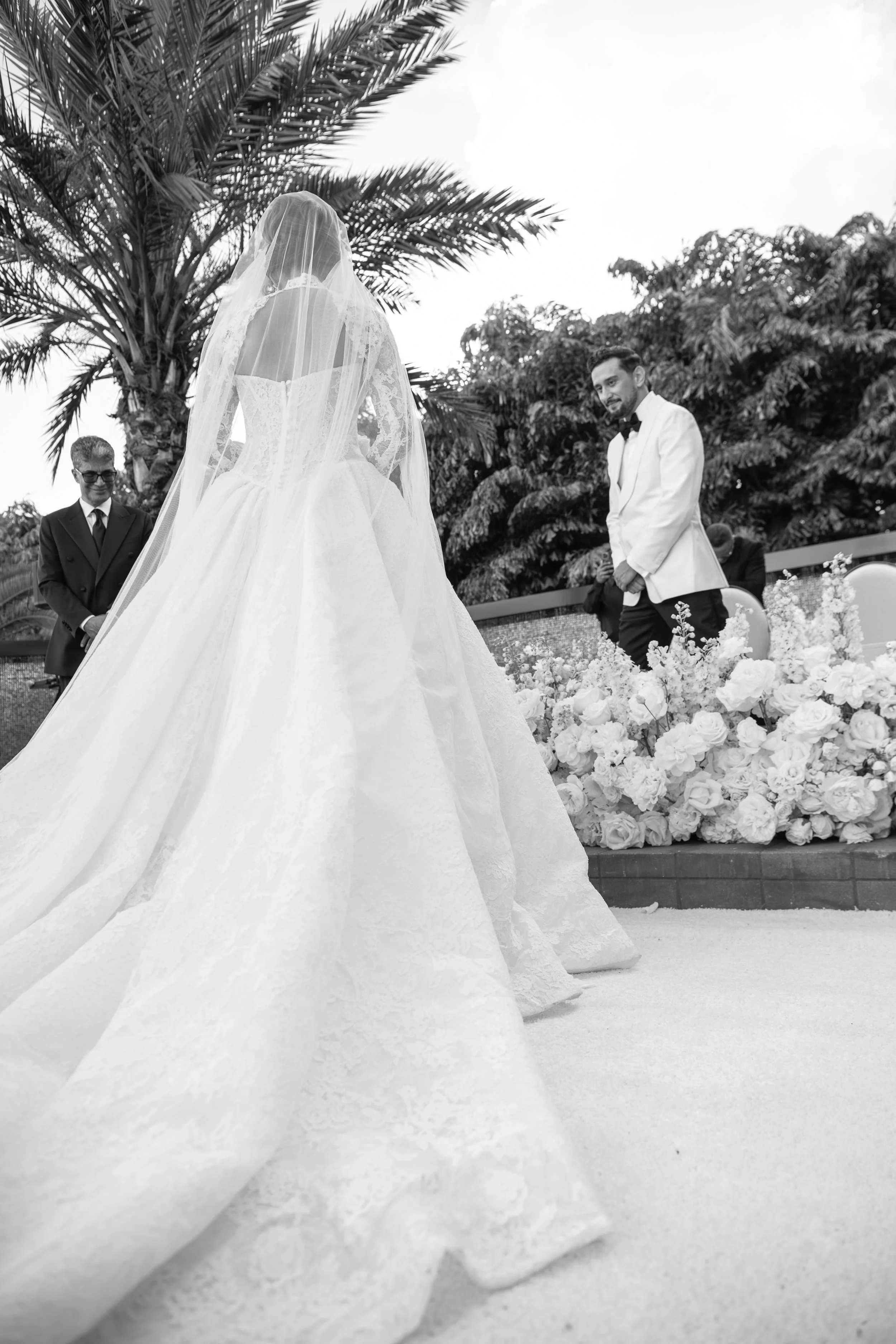Black and white photo of a wedding ceremony outdoors with a bride in a lace wedding gown and veil, groom in a tuxedo with bowtie, officiant, and floral decorations, surrounded by trees and palm fronds.