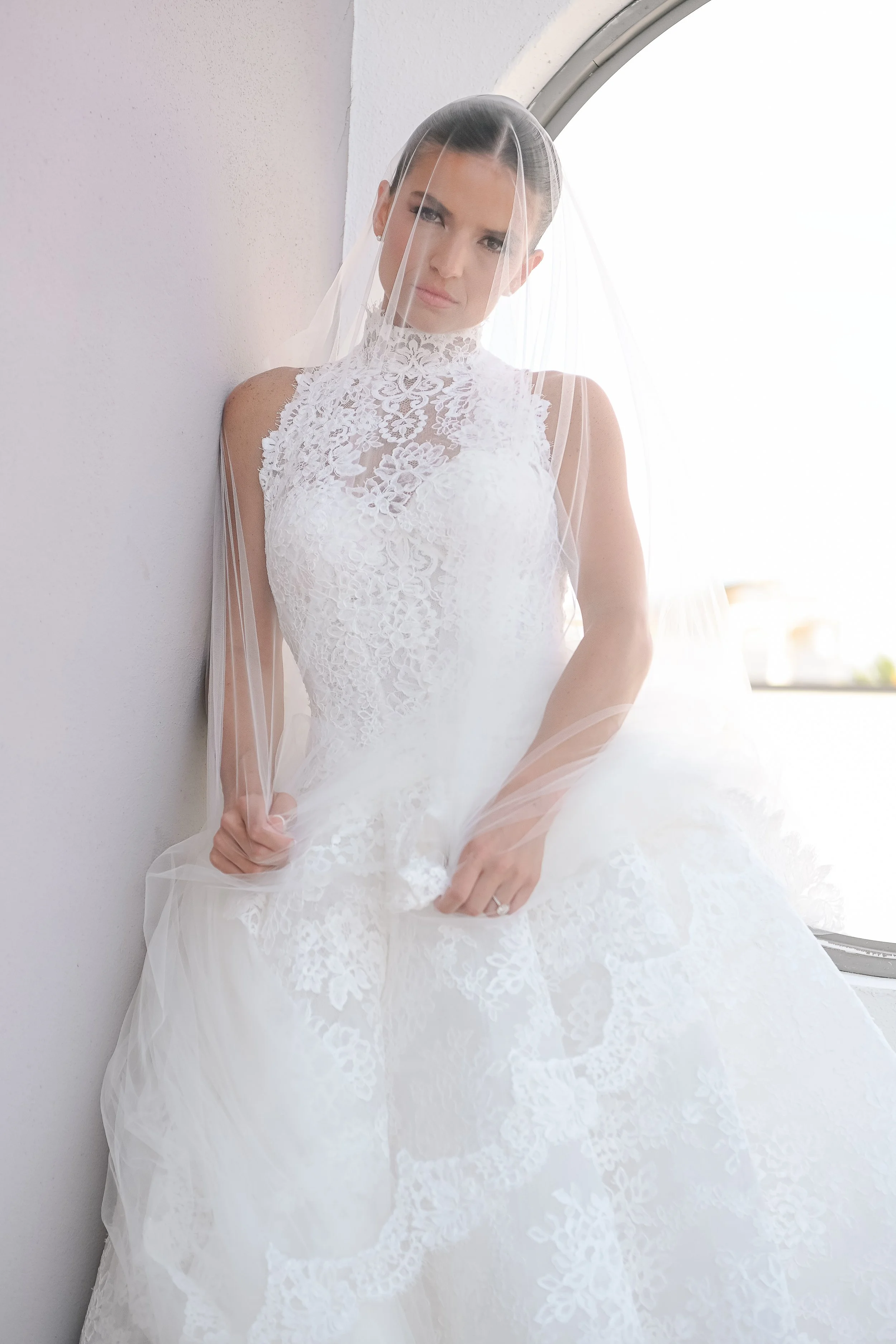 A woman in a white wedding dress with lace details and a veil, standing by a wall near a window, with natural light.