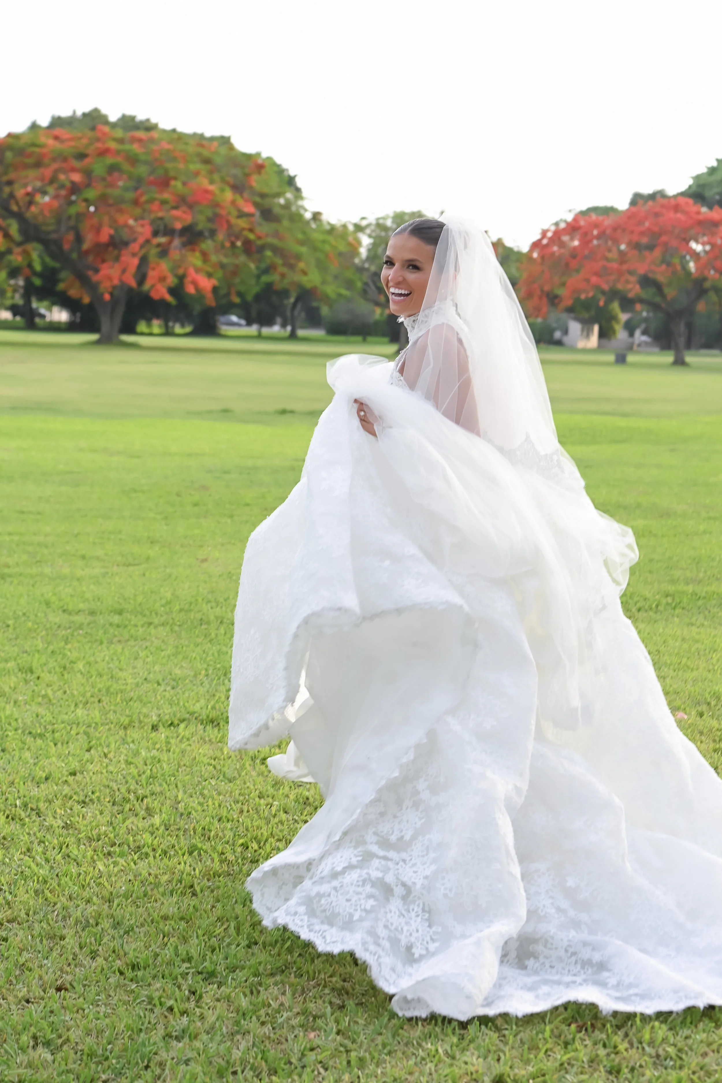 A smiling bride wearing a white wedding dress and veil, standing on a grassy field with trees that have red and orange leaves, suggesting an autumn setting.