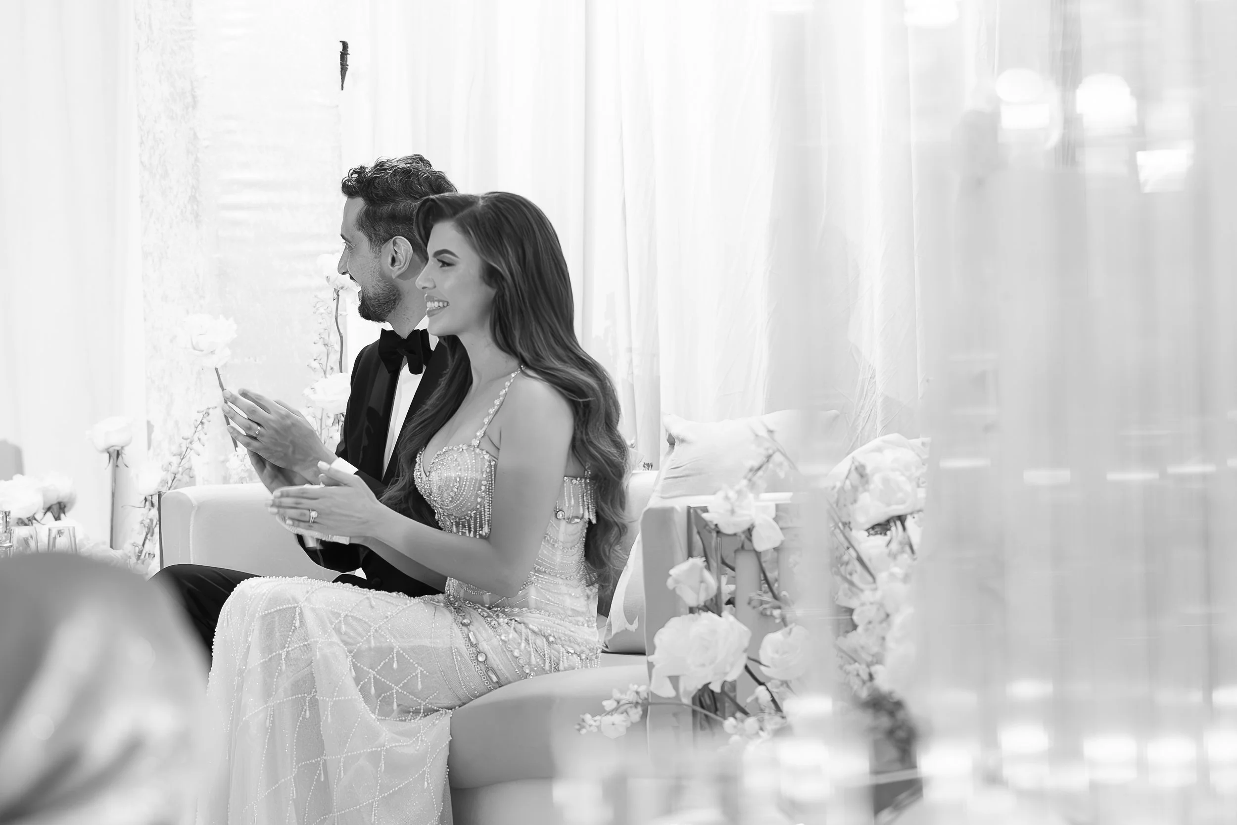 A bride and groom sit side by side during their wedding ceremony, smiling and looking forward, surrounded by floral decorations.