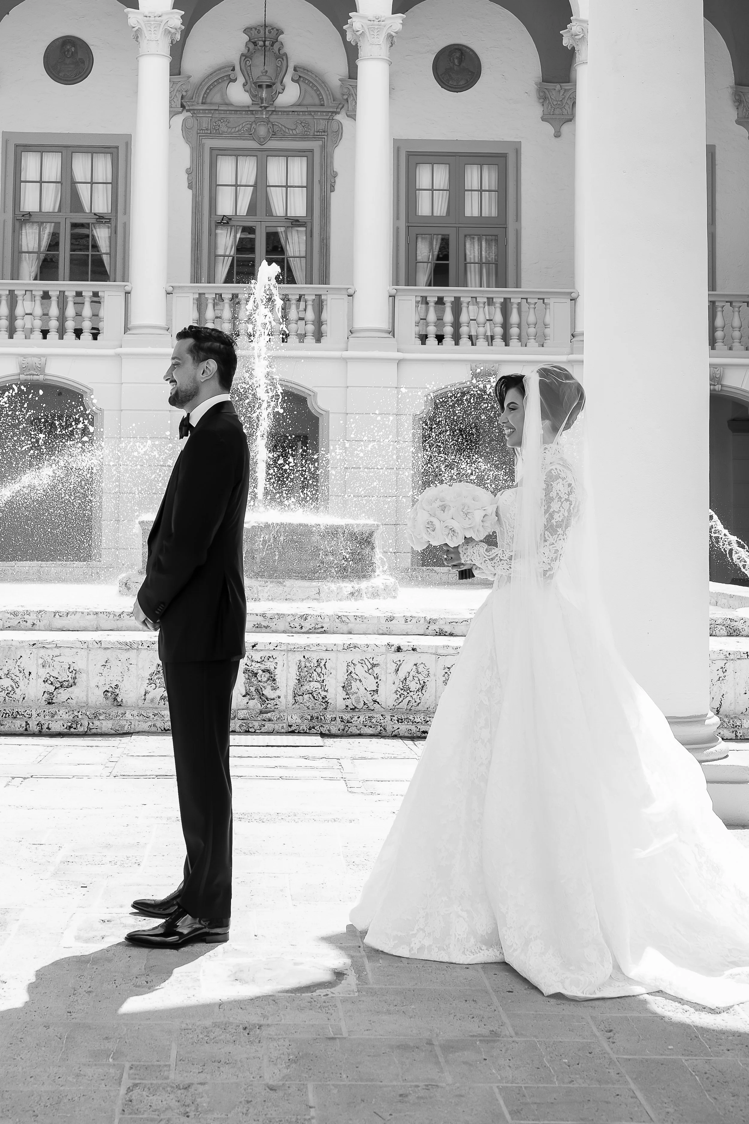 Black and white photo of a bride and groom standing apart in front of a fountain, with a European-style building in the background. The groom is in a tuxedo, facing left, smiling. The bride is in a wedding dress holding a bouquet of flowers, smiling 