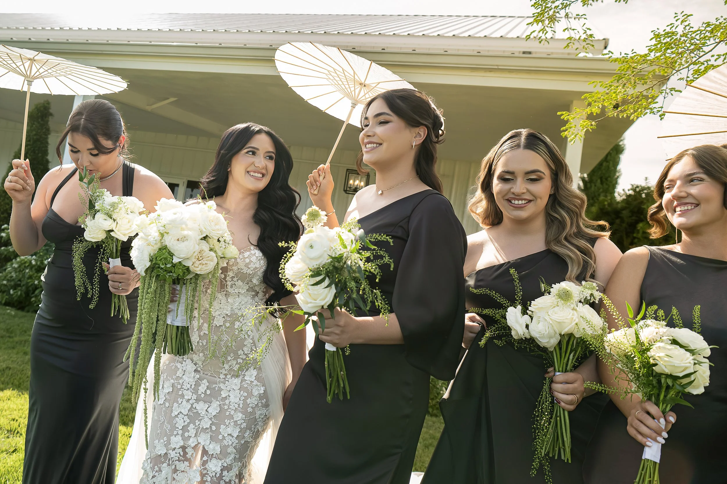 Group of women in black dresses holding white flower bouquets, standing under umbrellas outside during a wedding celebration.