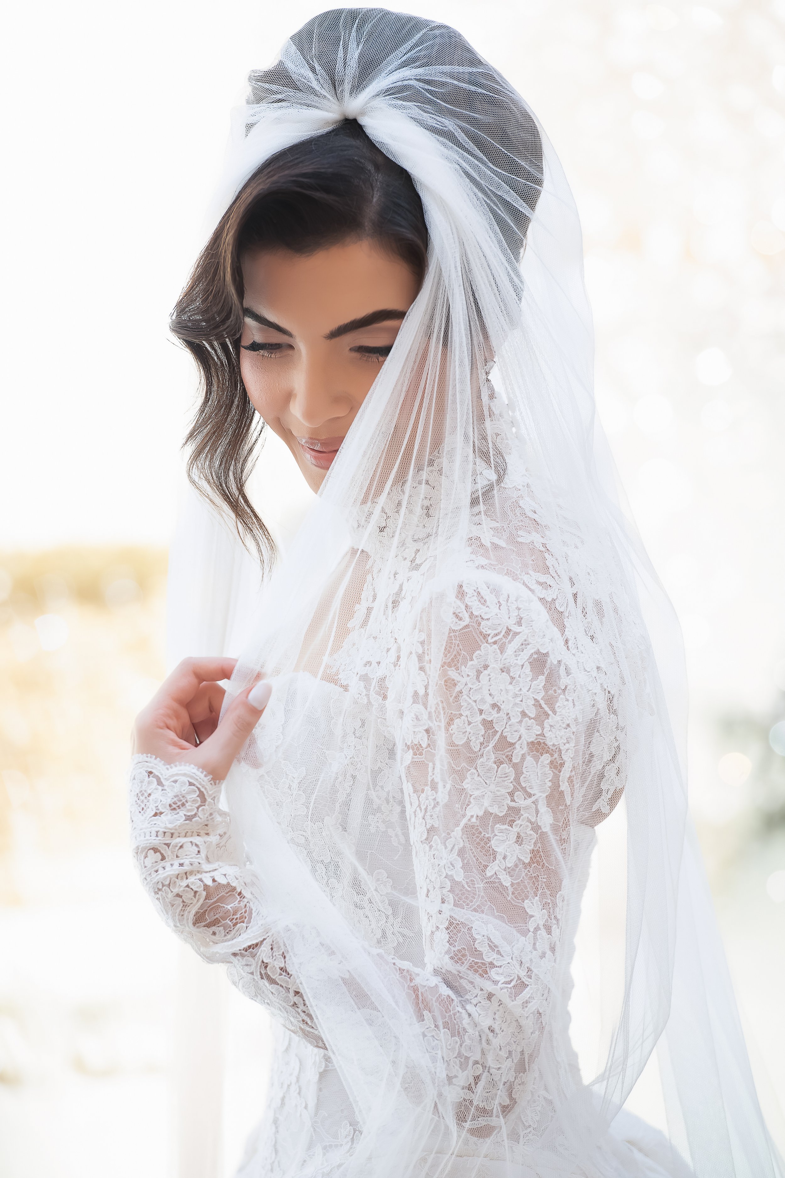 A woman dressed in a white lace wedding gown, with a sheer veil covering her dark, wavy hair and face, smiling softly outdoors.