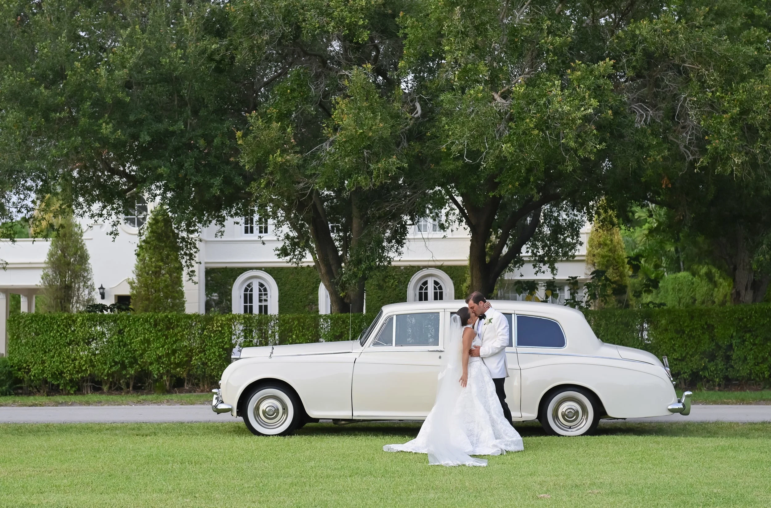 A bride and groom kissing next to a vintage white limousine in front of a large tree and a white building with arched windows.