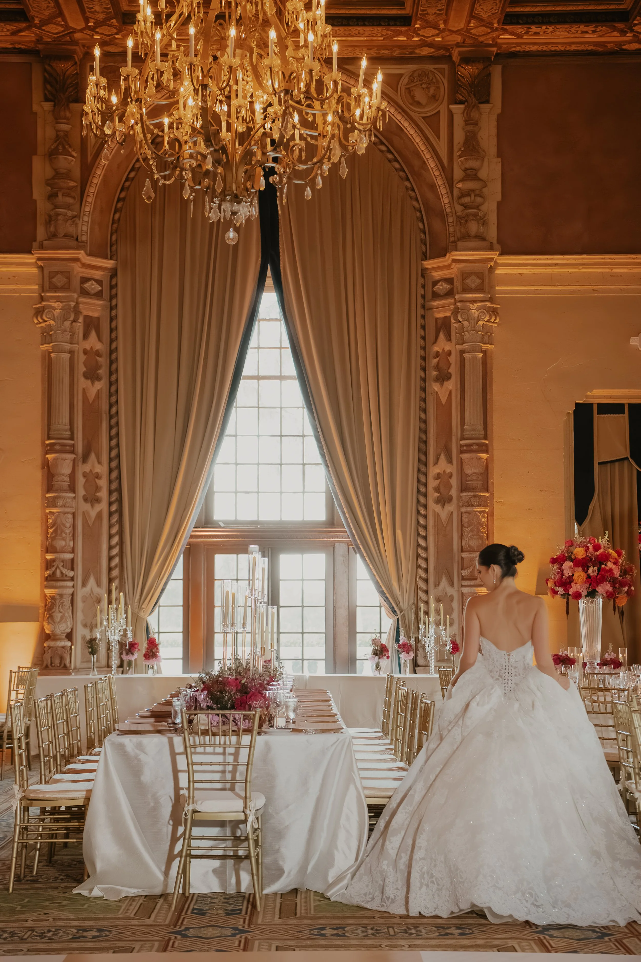 Elegant wedding reception with long banquet table covered in white tablecloths, gold chairs, and floral centerpieces. Large window behind with heavy curtains, ornate chandelier hanging from ceiling, a woman in a white wedding gown standing near the t