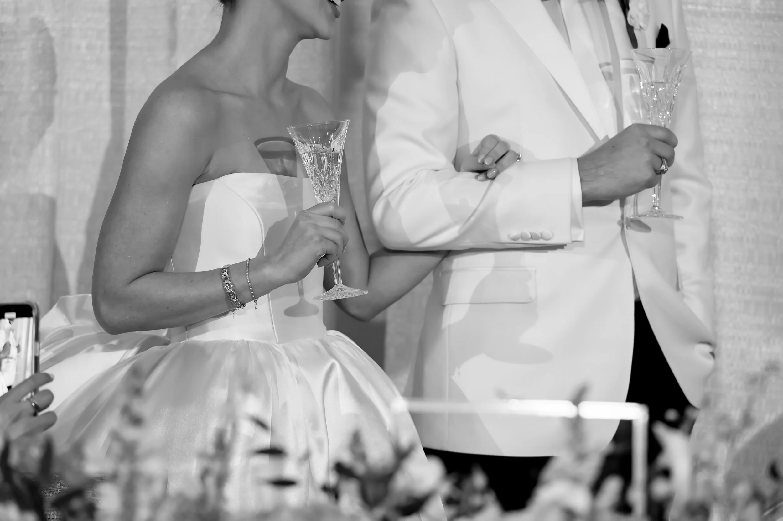 A bride and groom holding champagne glasses at a wedding reception. The bride wears a strapless wedding dress and a bracelet, the groom wears a white suit jacket. The background shows a textured wall.