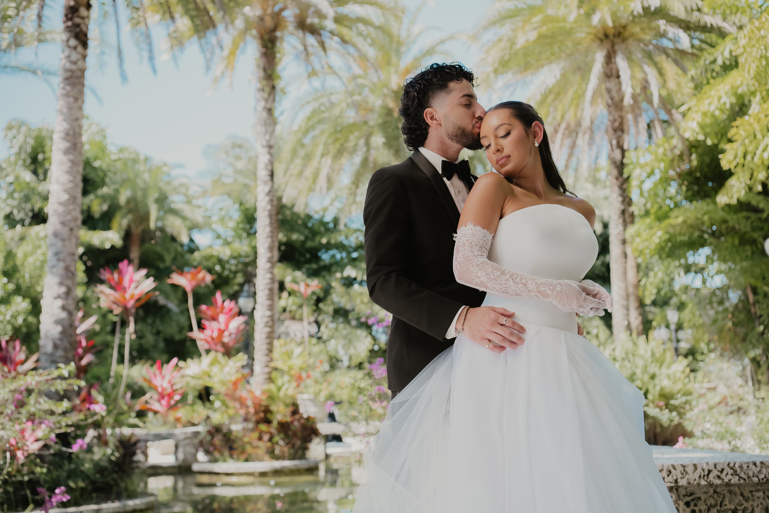 A groom in a black tuxedo and bow tie kisses a bride in a white wedding dress and long lace sleeves on her left arm, outdoors with palm trees and colorful flowers in the background.