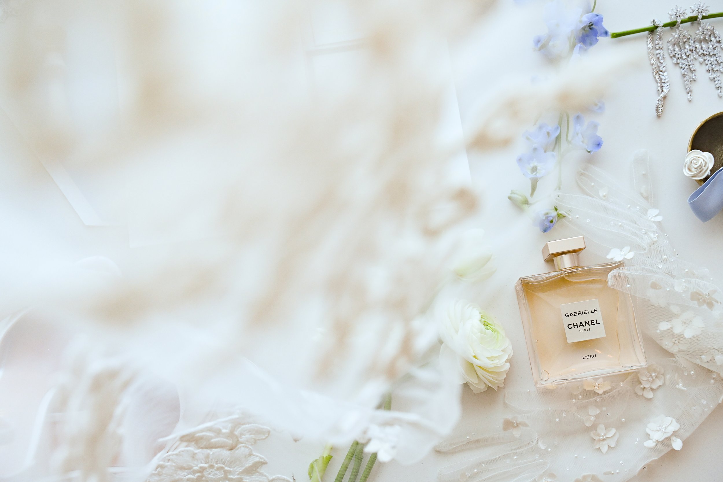 A flat lay of a white surface with a few light purple flowers, jewelry, and perfume bottles, including a Chanel perfume bottle labeled "Gabrielle".