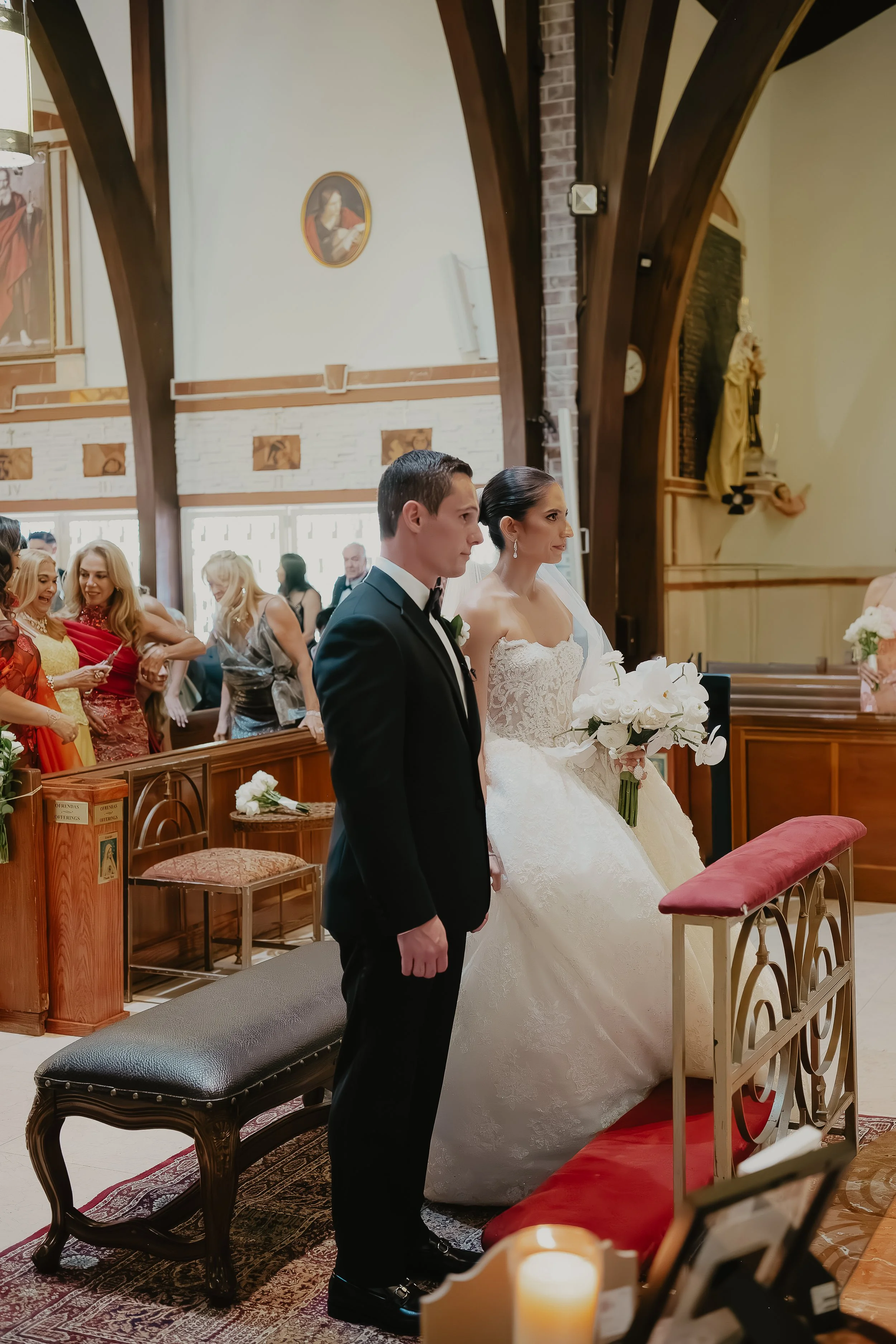 A wedding ceremony inside a church with a bride and groom standing side by side, with guests seated and standing behind them. The bride is in a white gown holding a bouquet, and the groom in a tuxedo. The church has wooden beams, religious artwork, a
