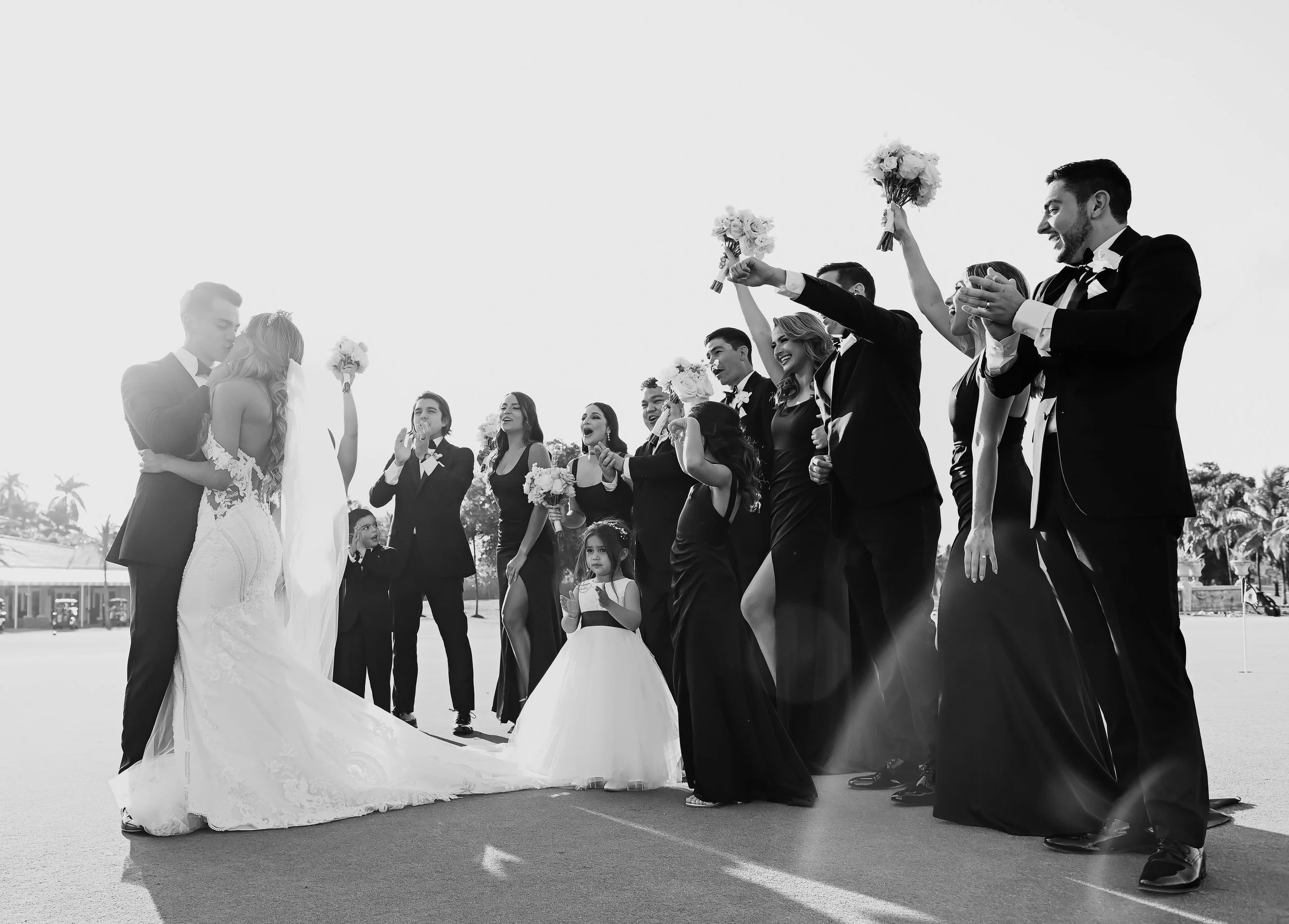 Black and white photo of a wedding scene with the bride and groom kissing, surrounded by other people holding bouquets and celebrating outdoors.