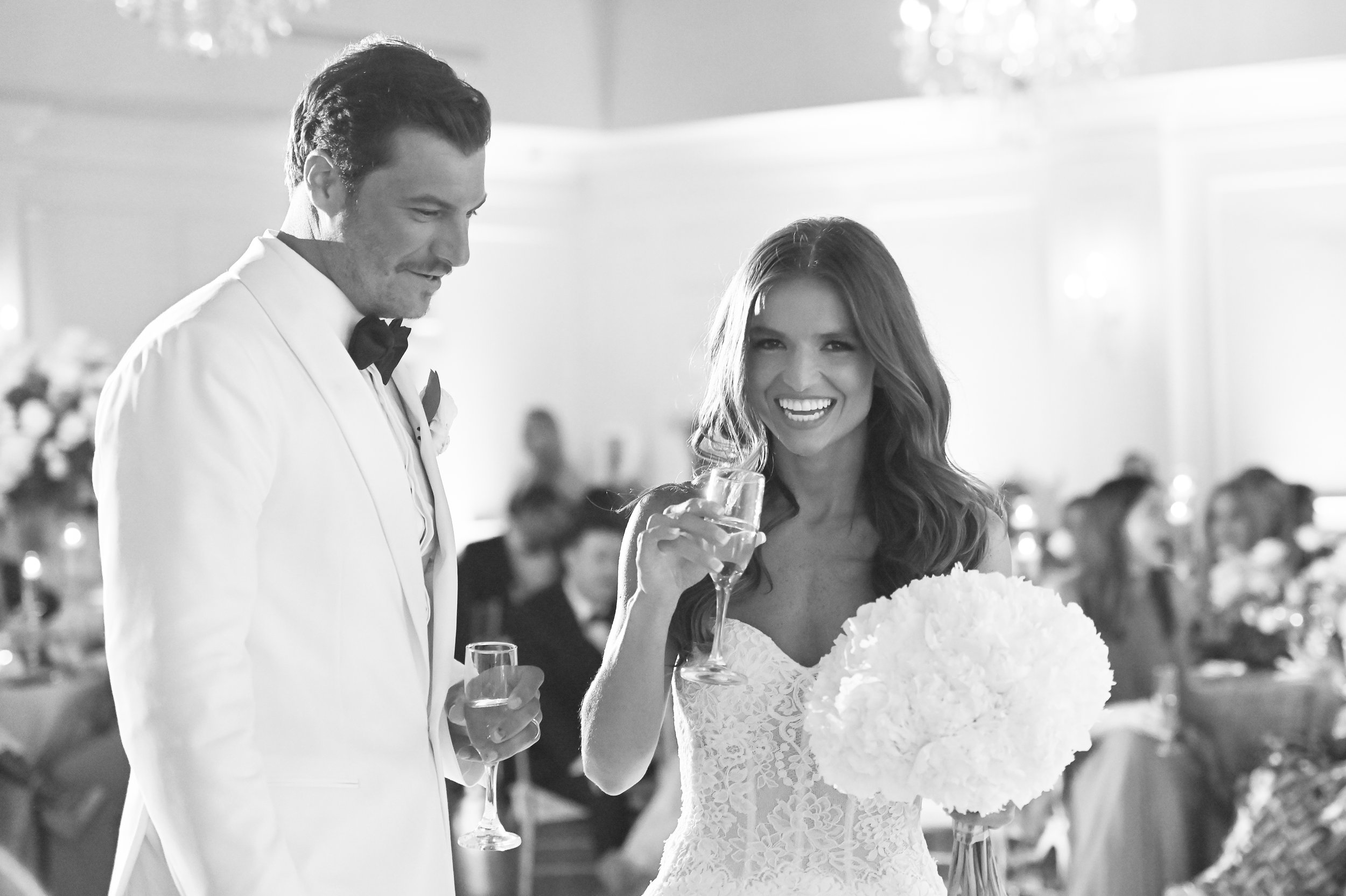 A bride and groom at a wedding reception, with the bride holding a glass of champagne and smiling, and the groom in a white tuxedo looking down.