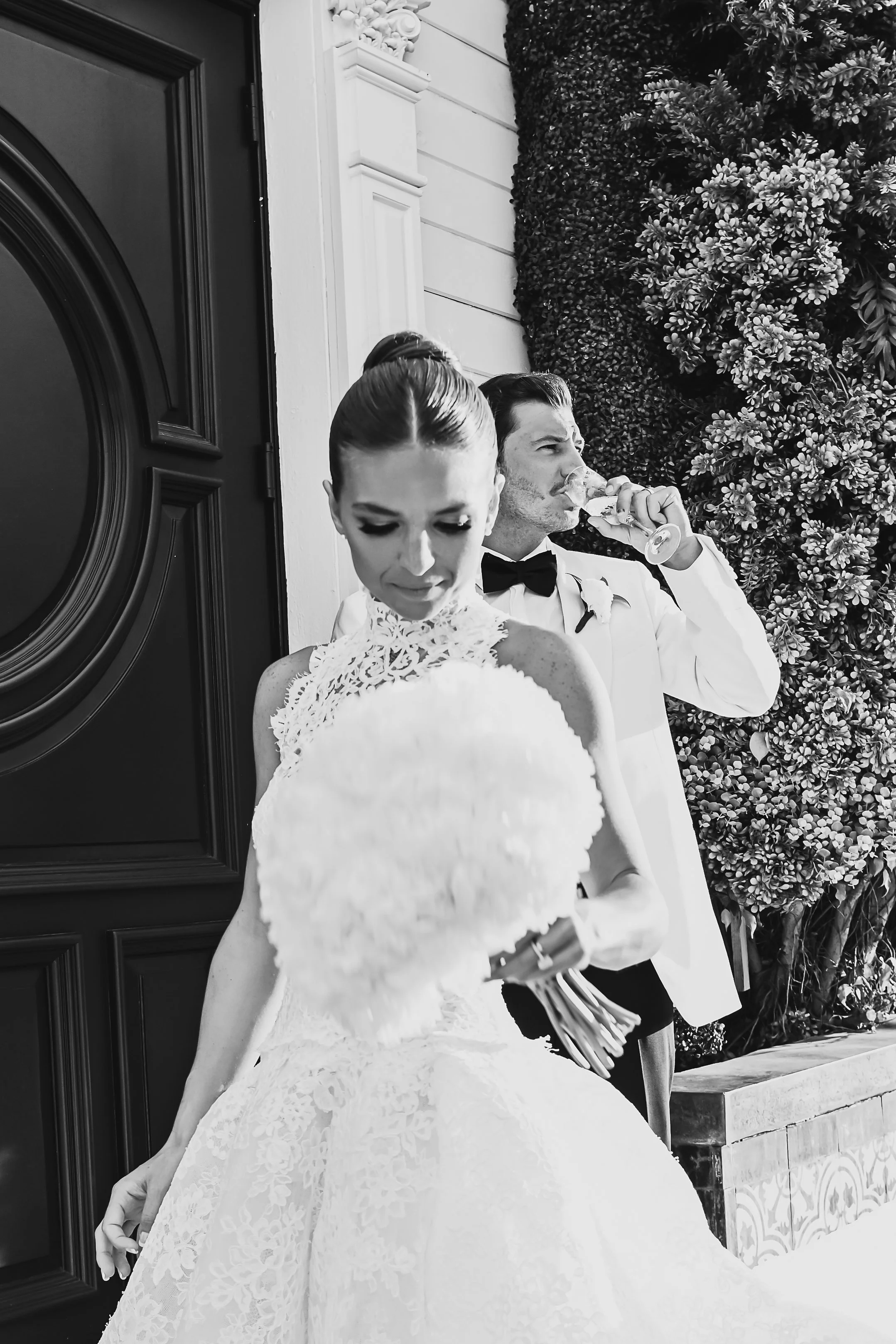 Black and white photo of a bride and groom standing outside a building. The bride wears a lace wedding dress and holds a bouquet, the groom in a white tuxedo with a black bow tie, drinking from a glass. There is a textured wall of greenery behind the