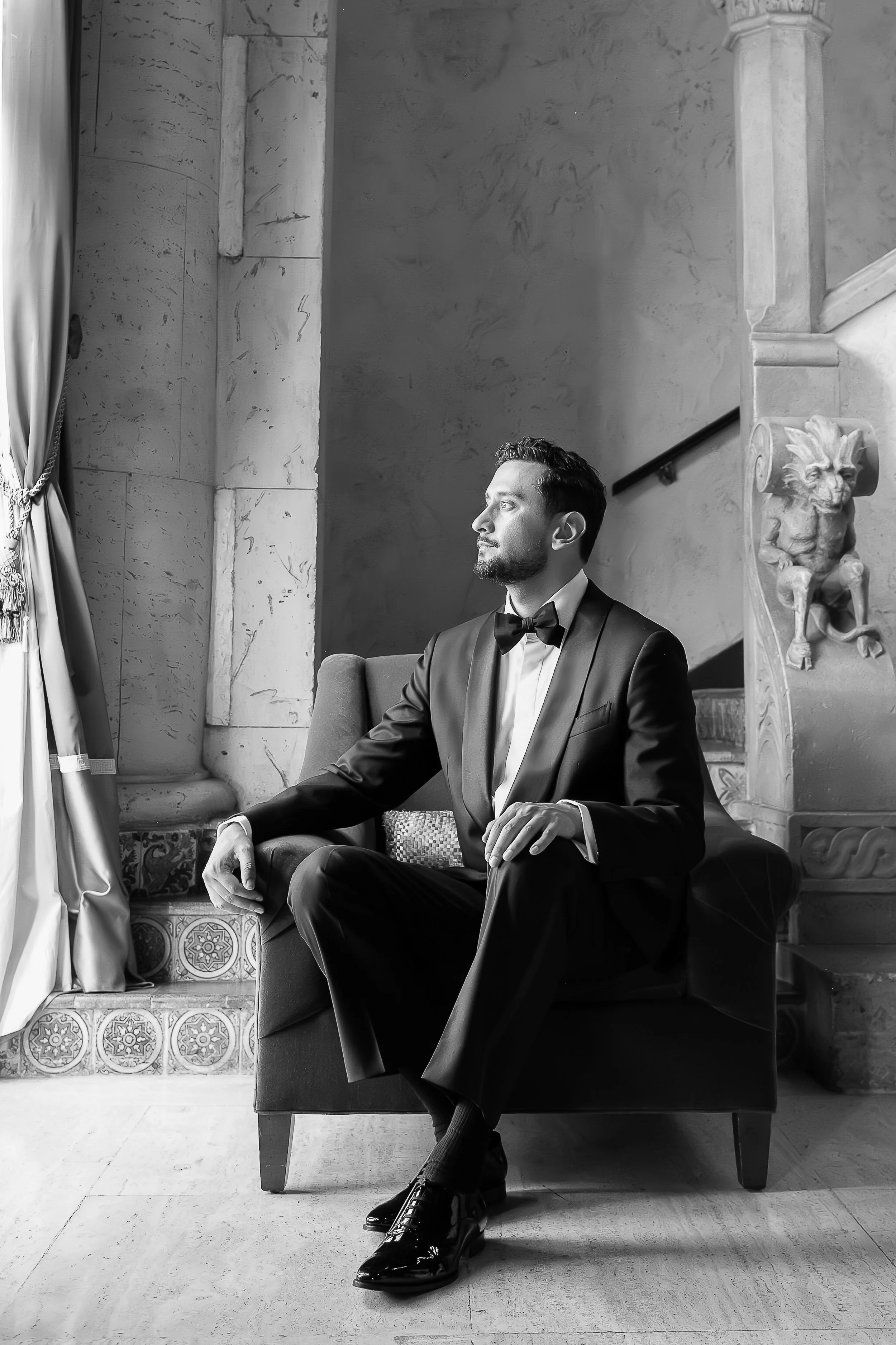 A man in a tuxedo sitting on a chair in a grand, ornate interior with stone columns and decorative elements, looking to the left.