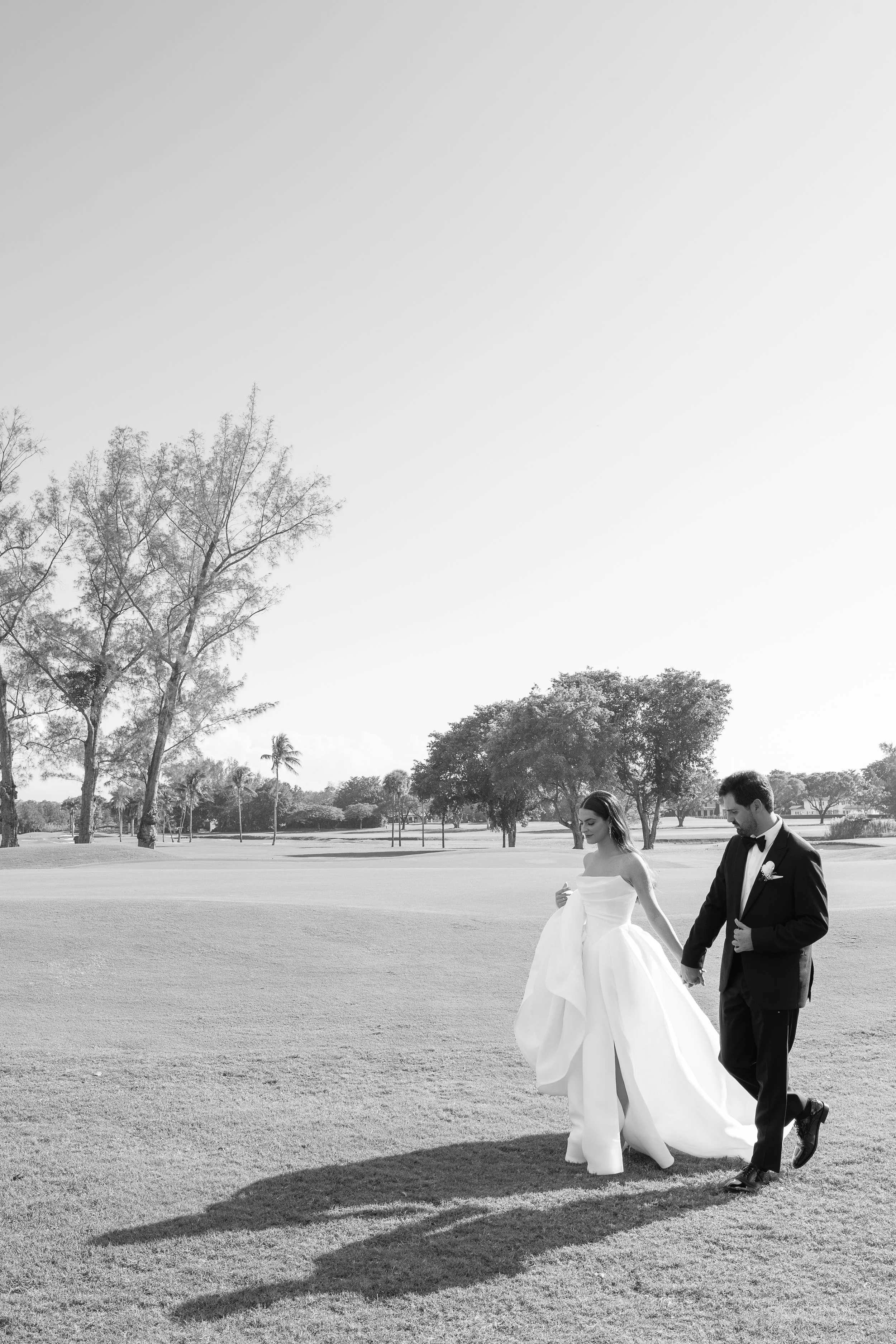 A black-and-white photo of a bride and groom walking on a golf course, surrounded by trees and open space, with the sunny sky above.