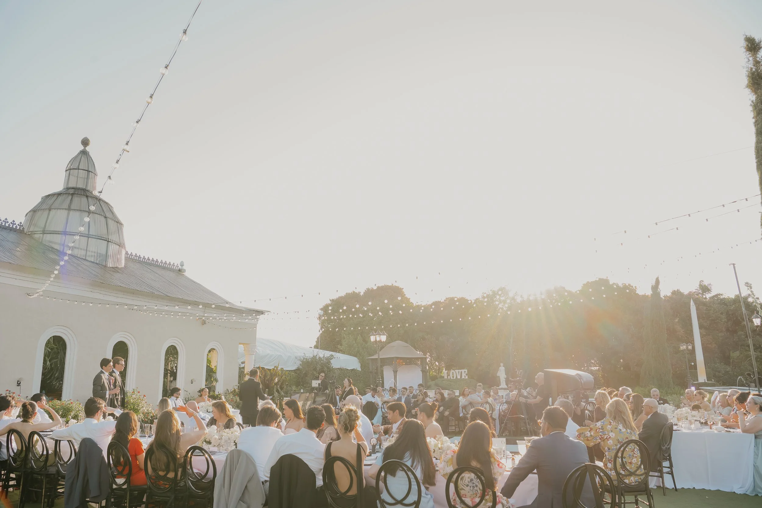 Outdoor wedding reception with guests seated at round tables, a white building with arched windows, string lights overhead, and a sunset background.