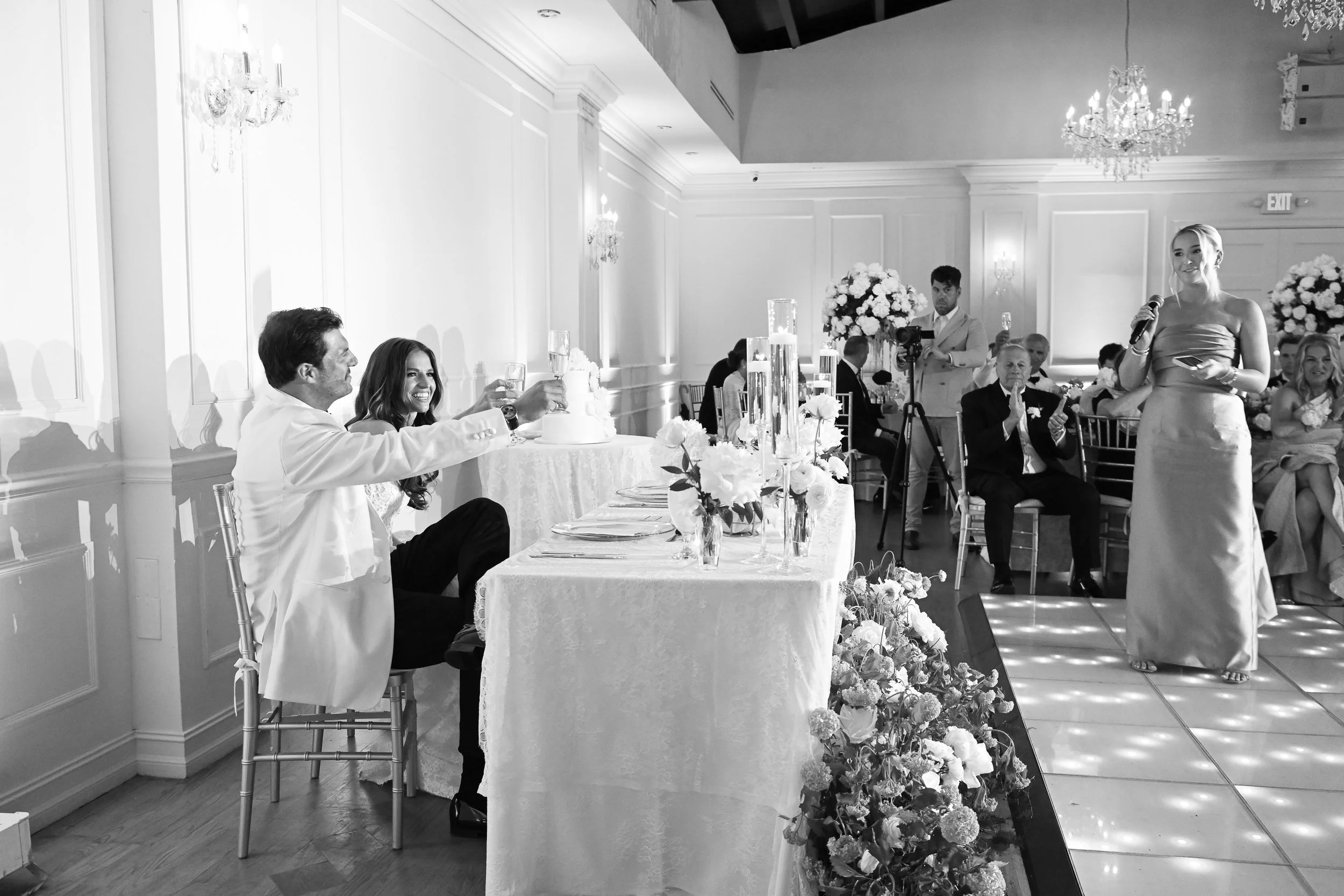 A wedding reception in an elegant banquet hall with white walls and chandeliers. The bride and groom are sitting at a table, smiling and raising glasses. A woman is giving a speech, holding a microphone. Guests are seated around, and floral arrangeme