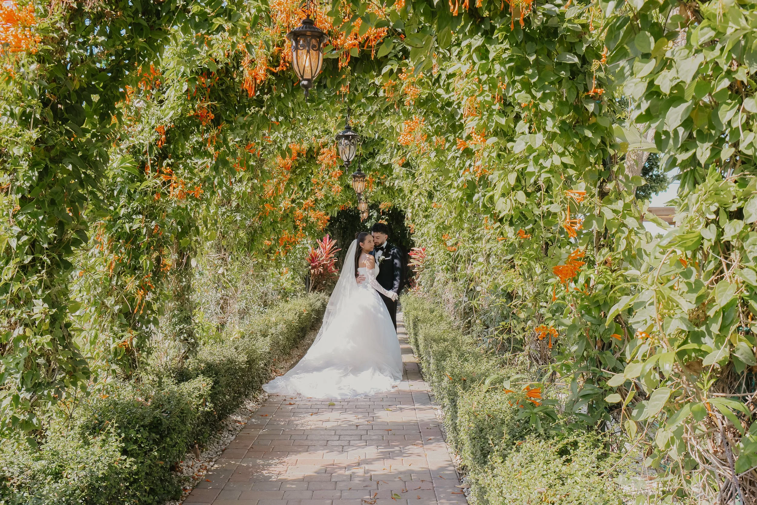 A bride and groom standing together in a lush, green garden aisle under a canopy of leafy vines and orange flowers, with hanging lanterns overhead.