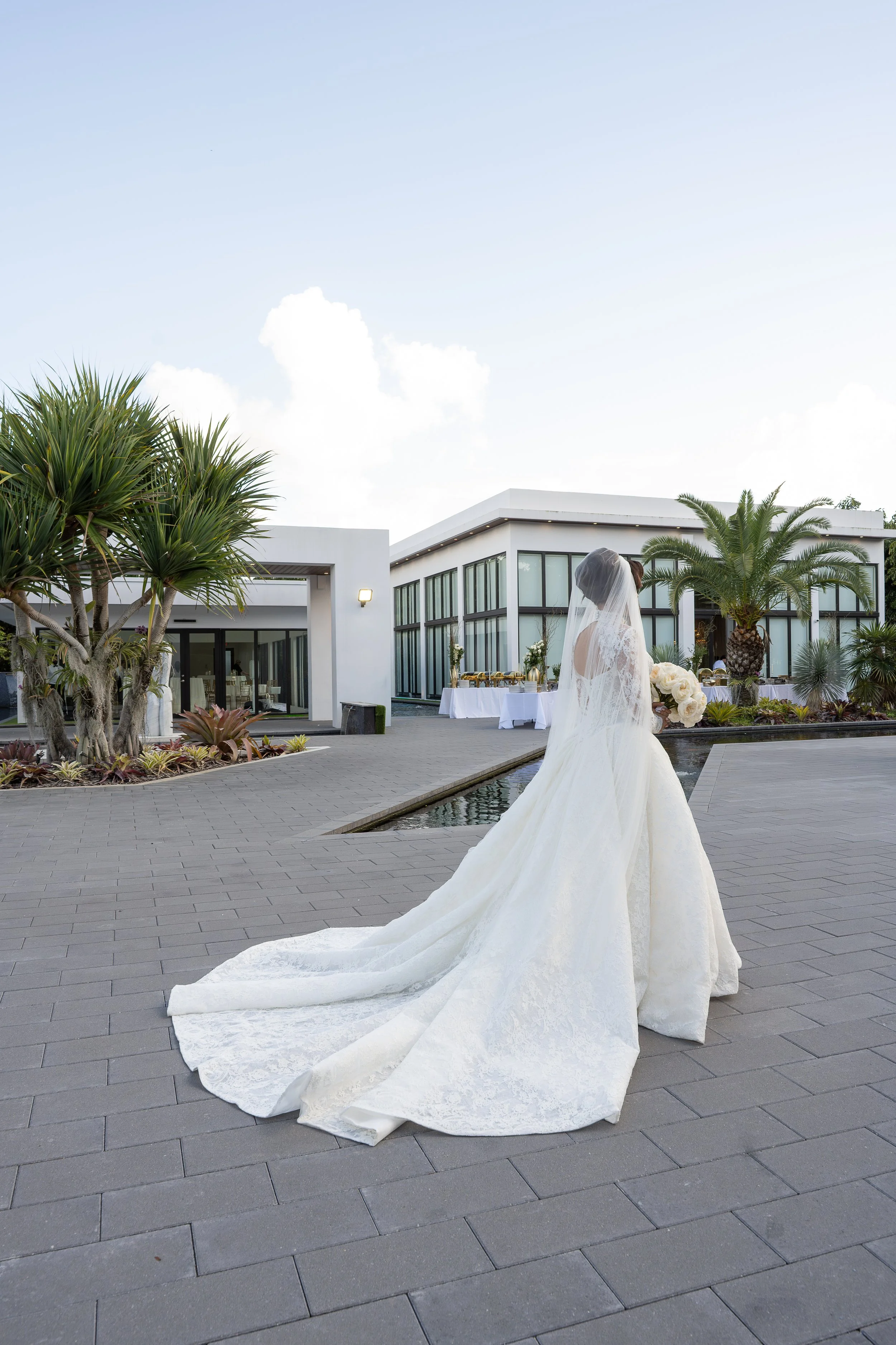 A bride in a white wedding dress and veil stands near a water feature outside a modern white building with large windows, surrounded by tropical plants and palm trees.