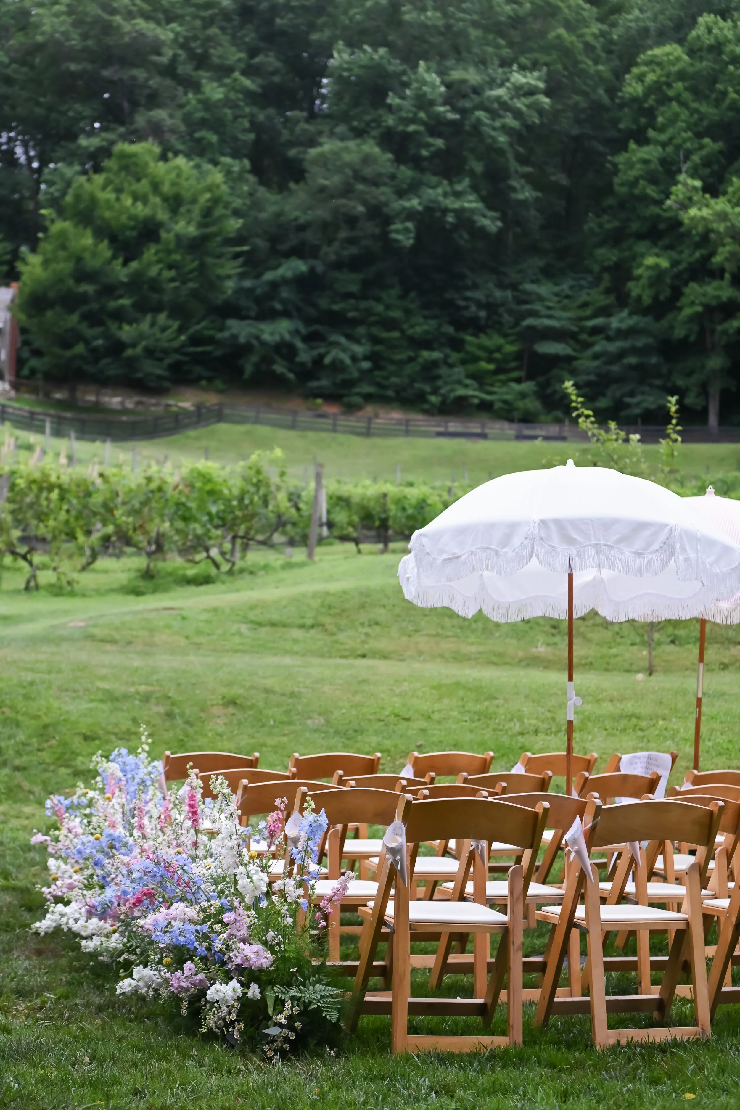 Outdoor wedding setup with wooden chairs, white umbrellas, and colorful flower arrangements on a lawn surrounded by trees.