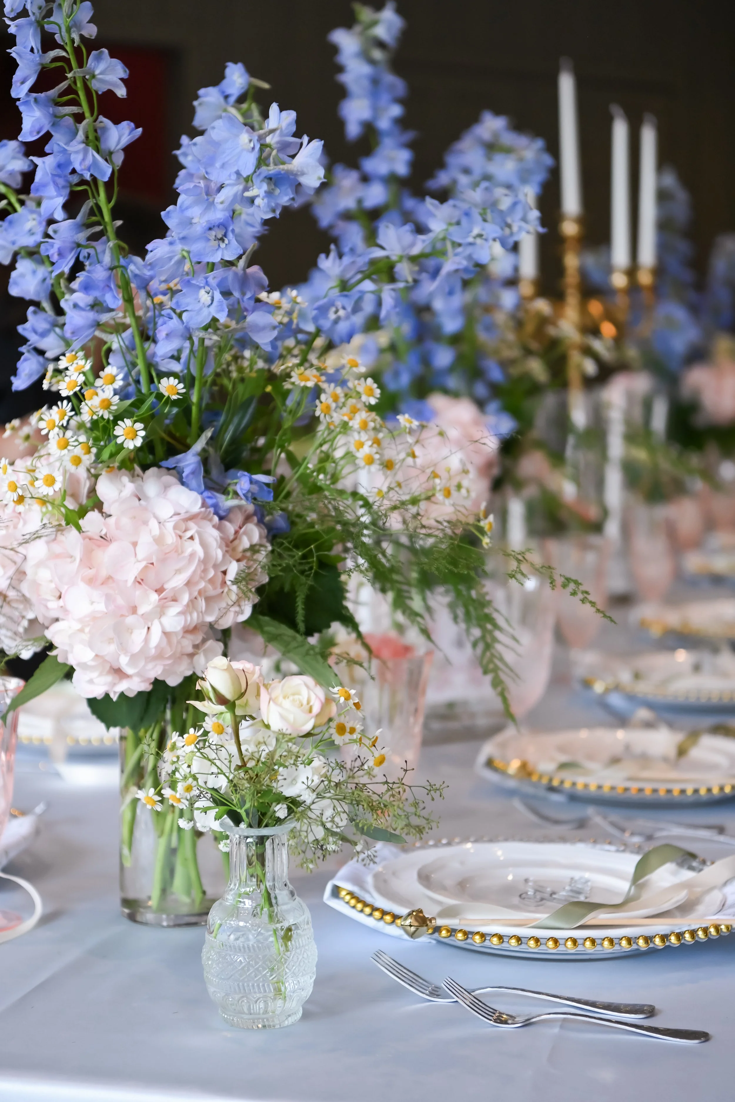A floral centerpiece with blue delphinium, pink hydrangeas, white daisies, and greenery on a white table setting with fine china and gold accents, set for a formal event.