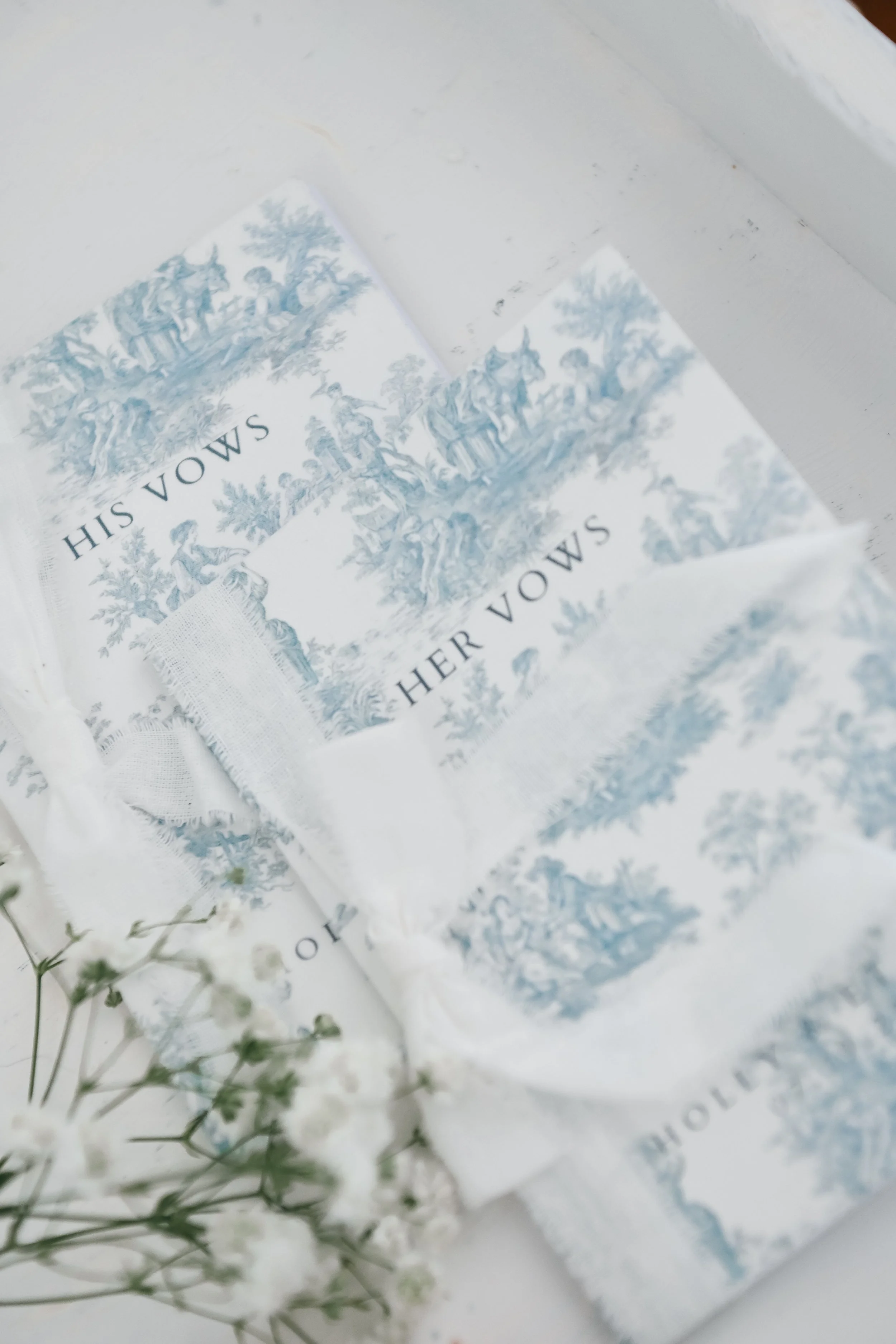 Close-up of a paper or card with a vintage-style blue and white illustration and the words 'His Vows' visible. Some blurred white flowers or plant stems are in the foreground.