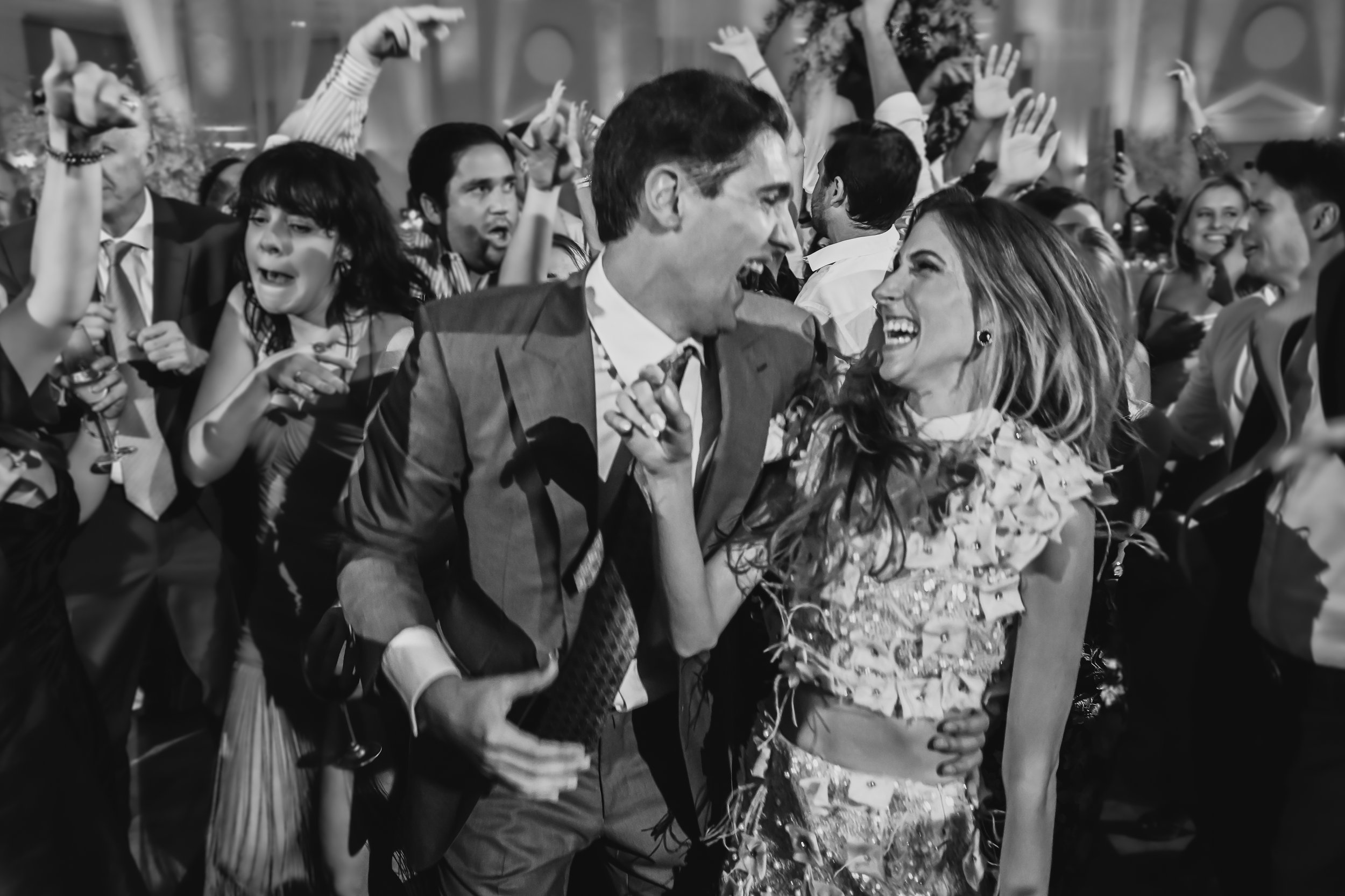 A lively celebration scene with people dressed in formal attire, dancing, and smiling at an indoor event.