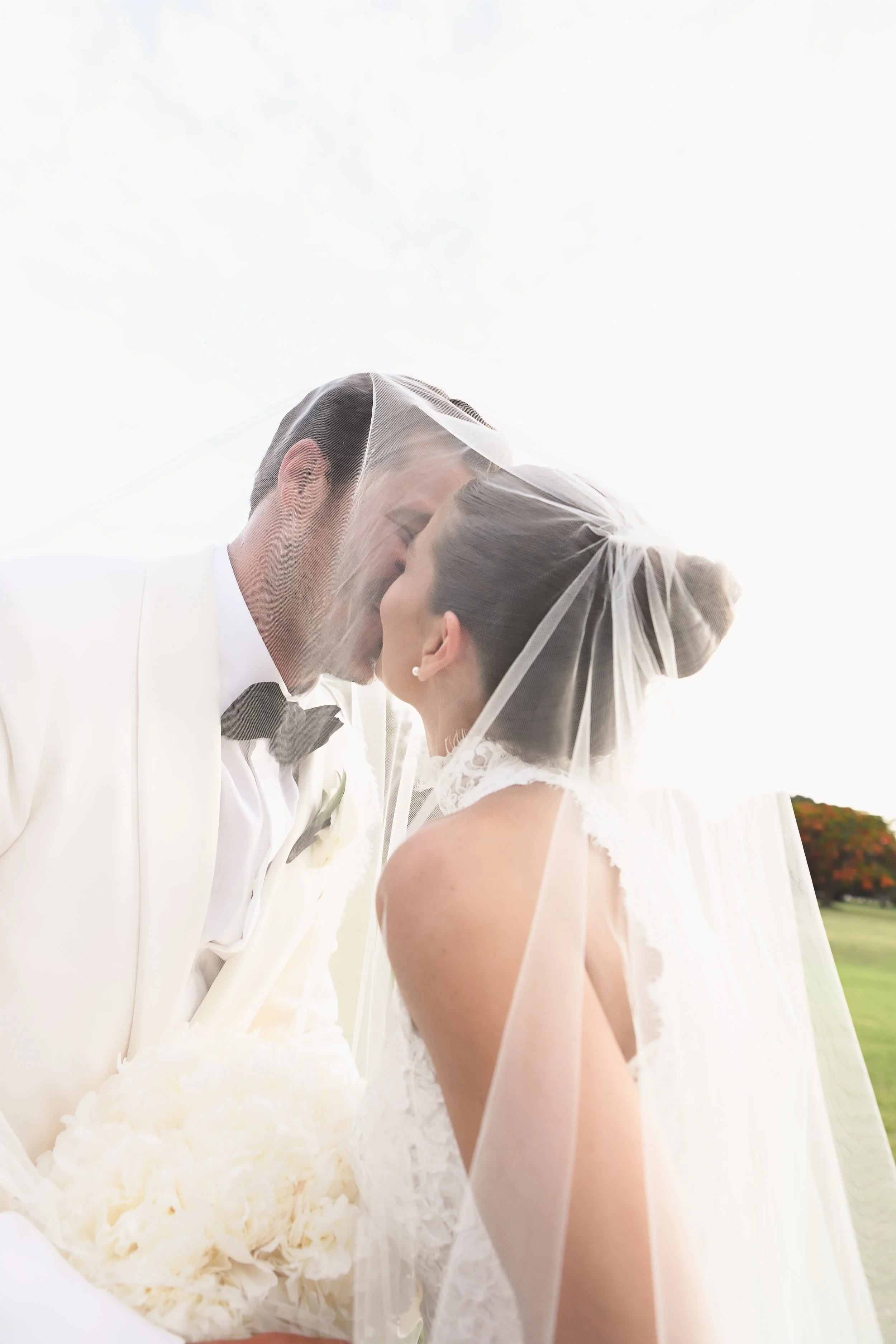 A bride and groom are close, touching foreheads, with the groom holding a bouquet of cream-colored flowers, both wearing wedding attire, outdoors on a bright day.