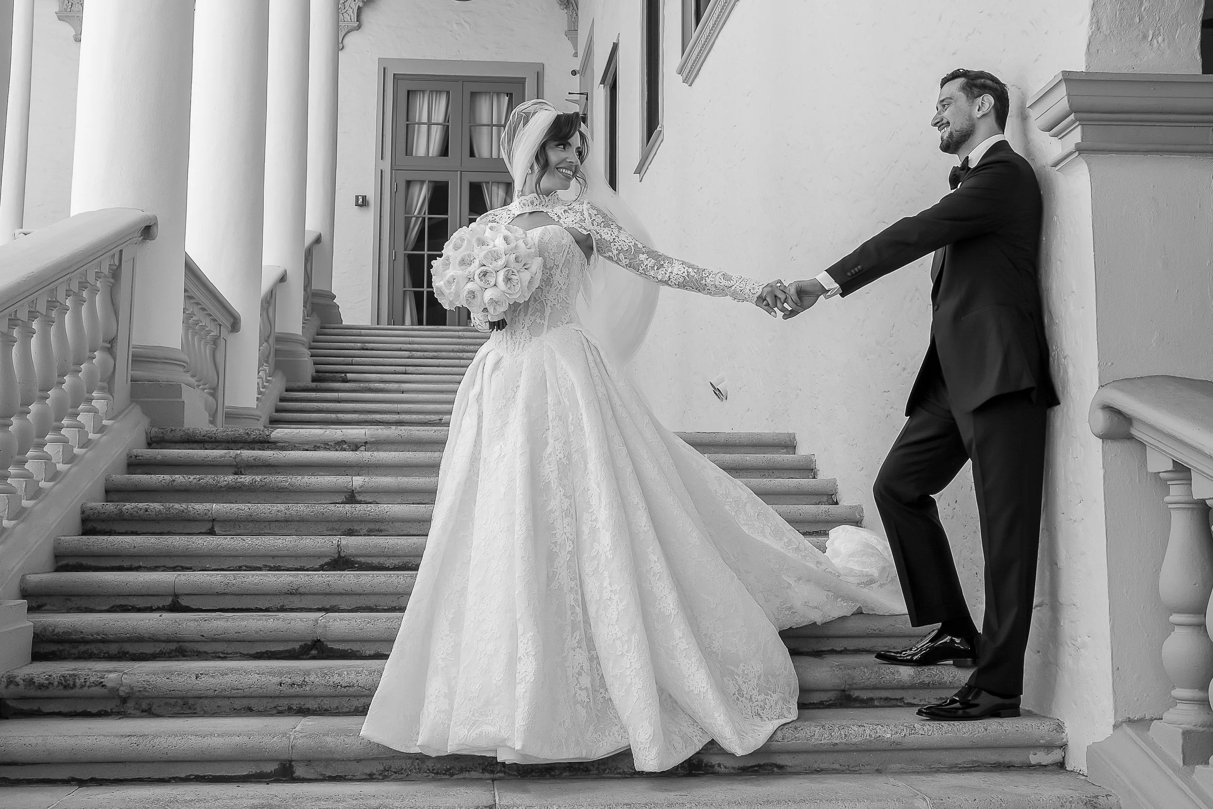 Wedding couple on stairs, the bride in a lace wedding gown holding a bouquet, and the groom in a black tuxedo, smiling at each other.