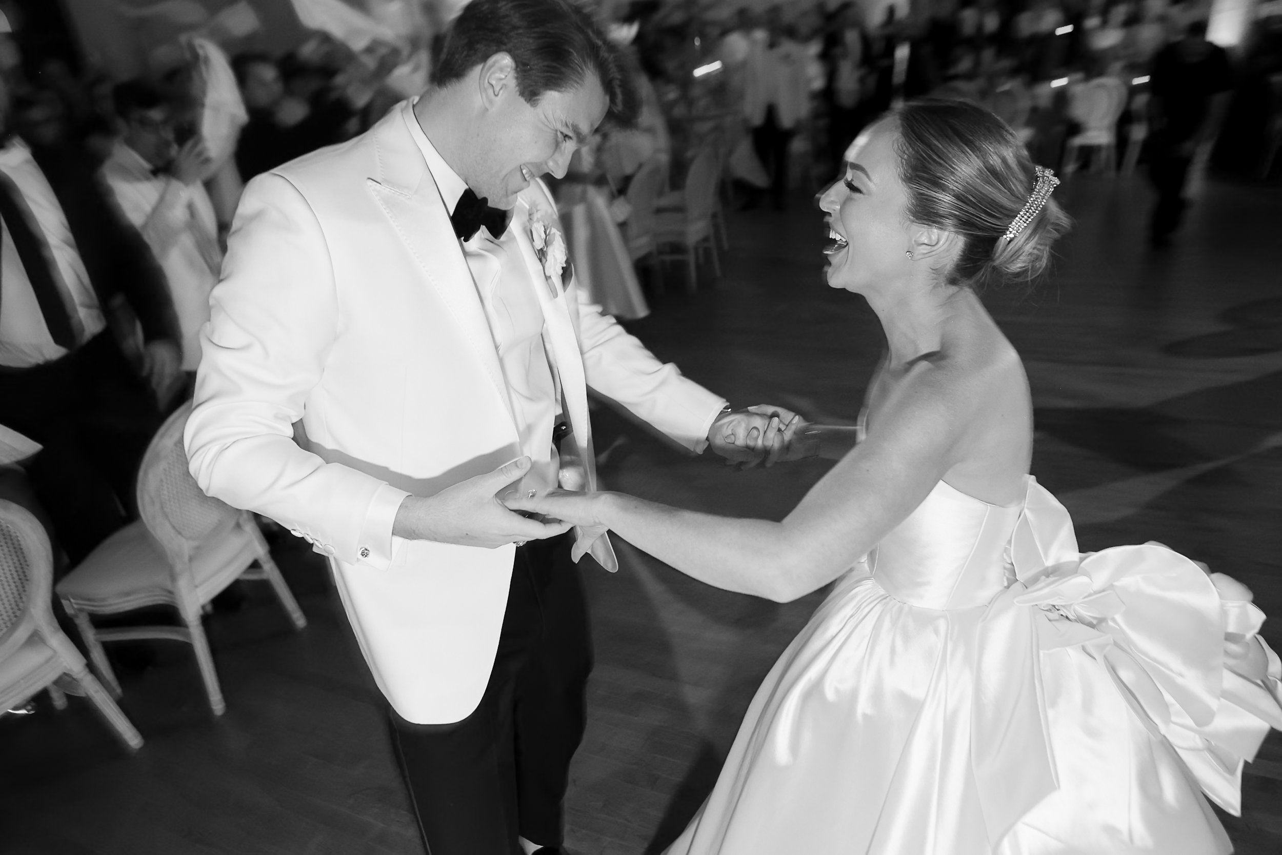 A bride and groom are dancing and laughing at their wedding reception. The groom is wearing a white tuxedo with a black bow tie, and the bride is wearing a strapless wedding gown with a pearl hair accessory.