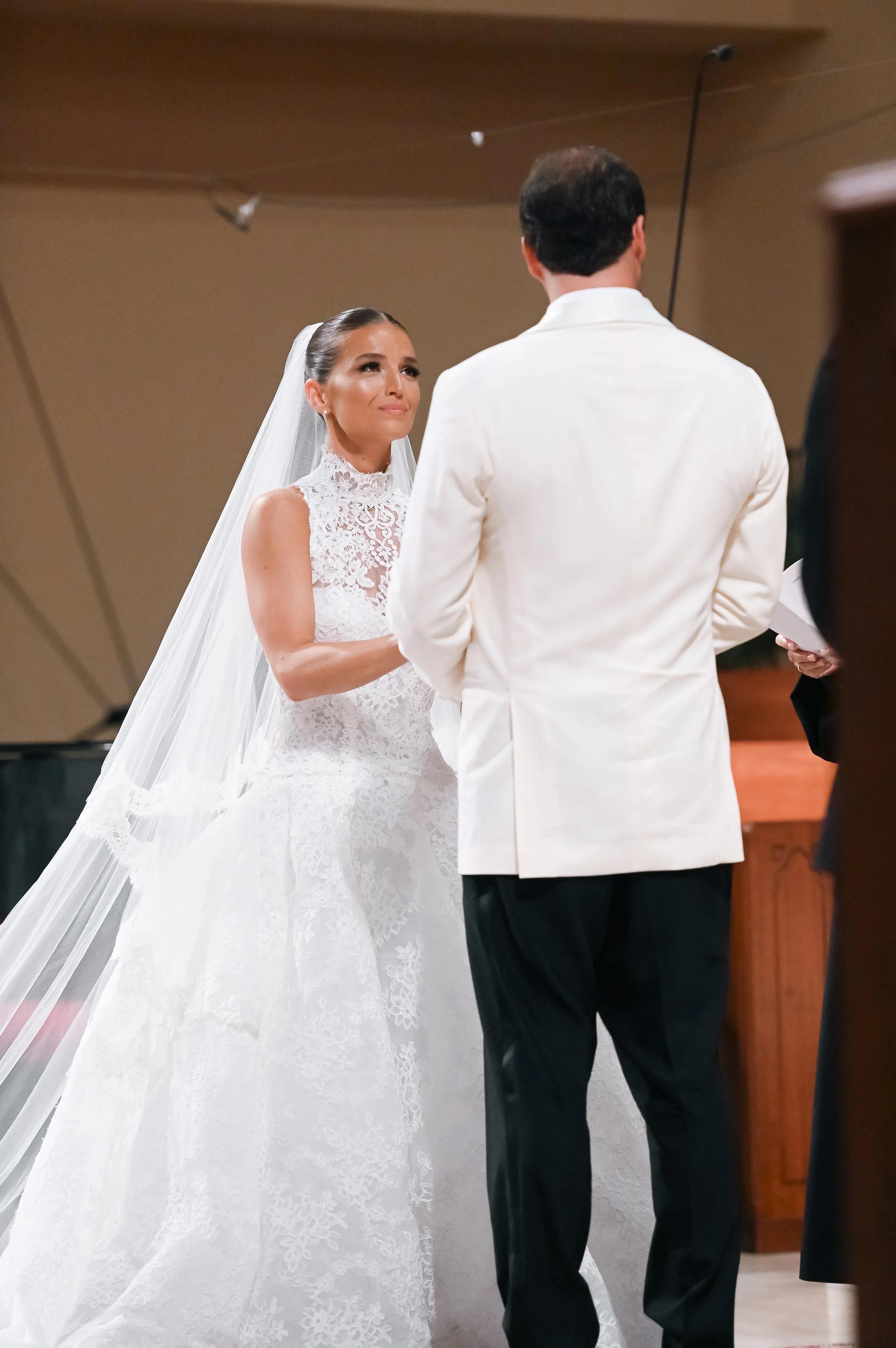 A bride wearing a white lace wedding gown with a long veil and a groom in a white jacket standing at the altar during their wedding ceremony.