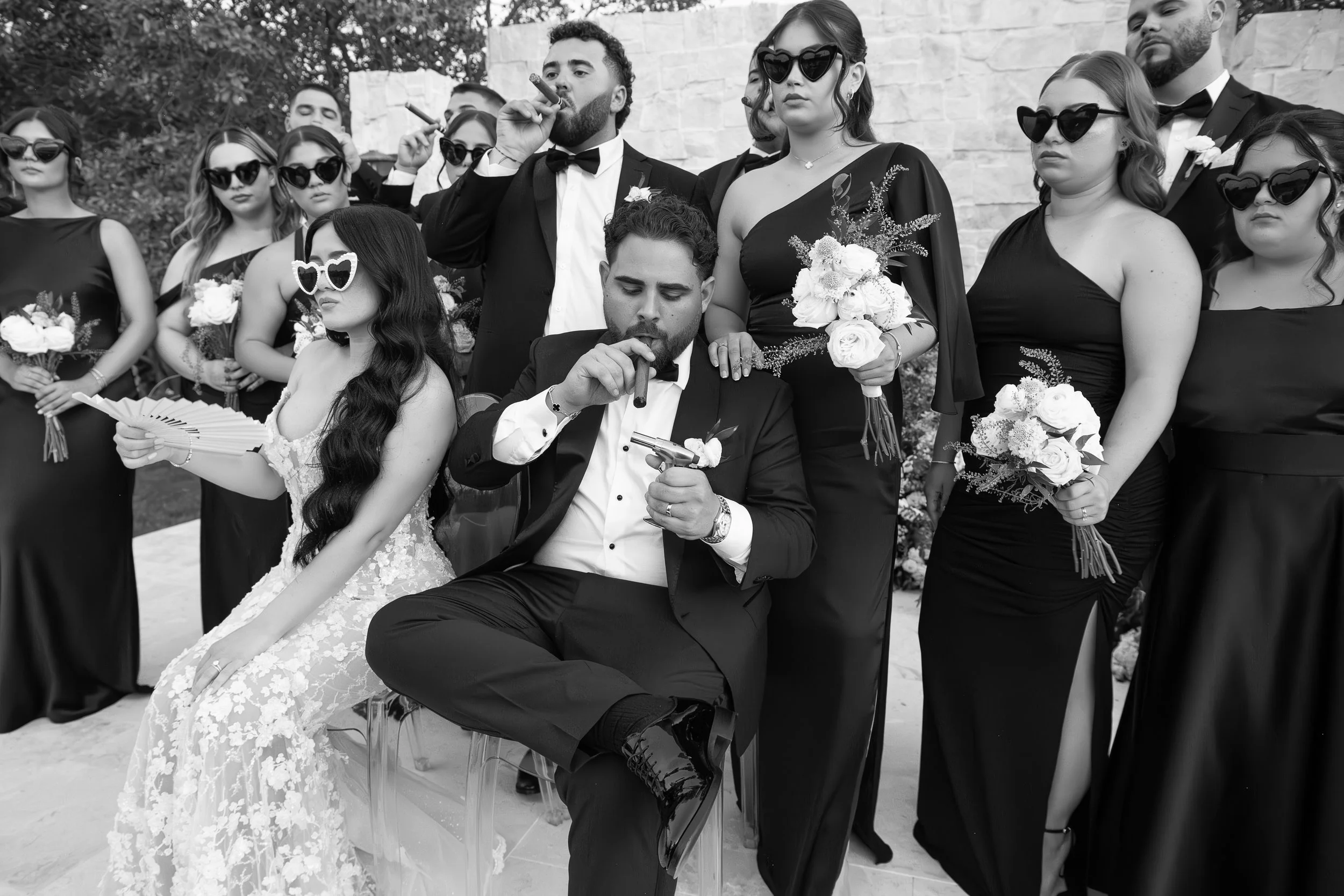 Group of people dressed in formal attire, with women in black dresses, some wearing sunglasses, and men in tuxedos, at an outdoor event. Three individuals in the front are smoking cigars, and a woman is holding a bouquet of flowers.