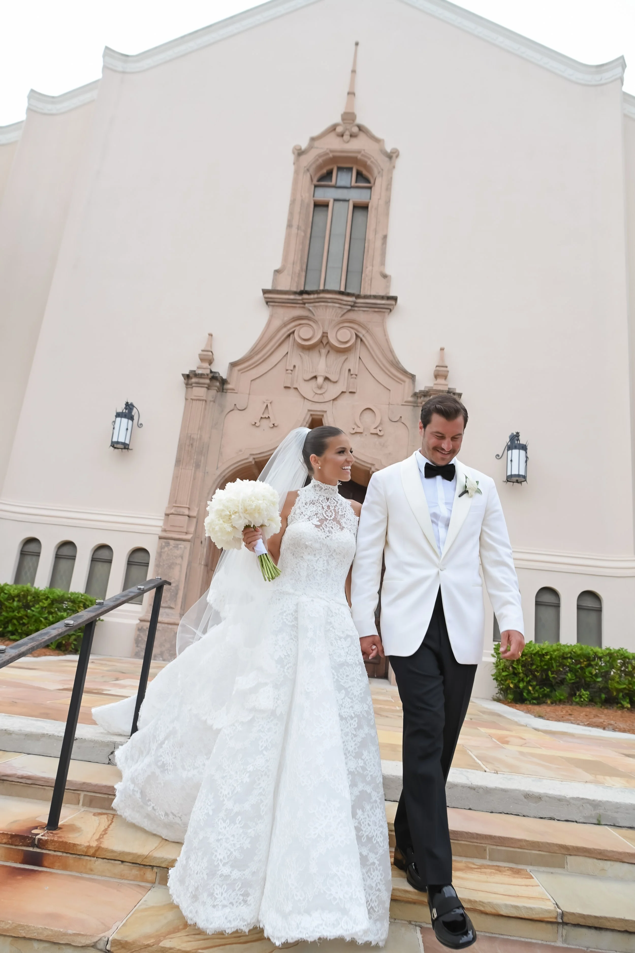 A bride and groom walking hand in hand outside a church building on their wedding day. The bride is wearing a lace wedding gown and holding a bouquet of white flowers, while the groom is dressed in a white tuxedo jacket with black pants and a black bow tie.
