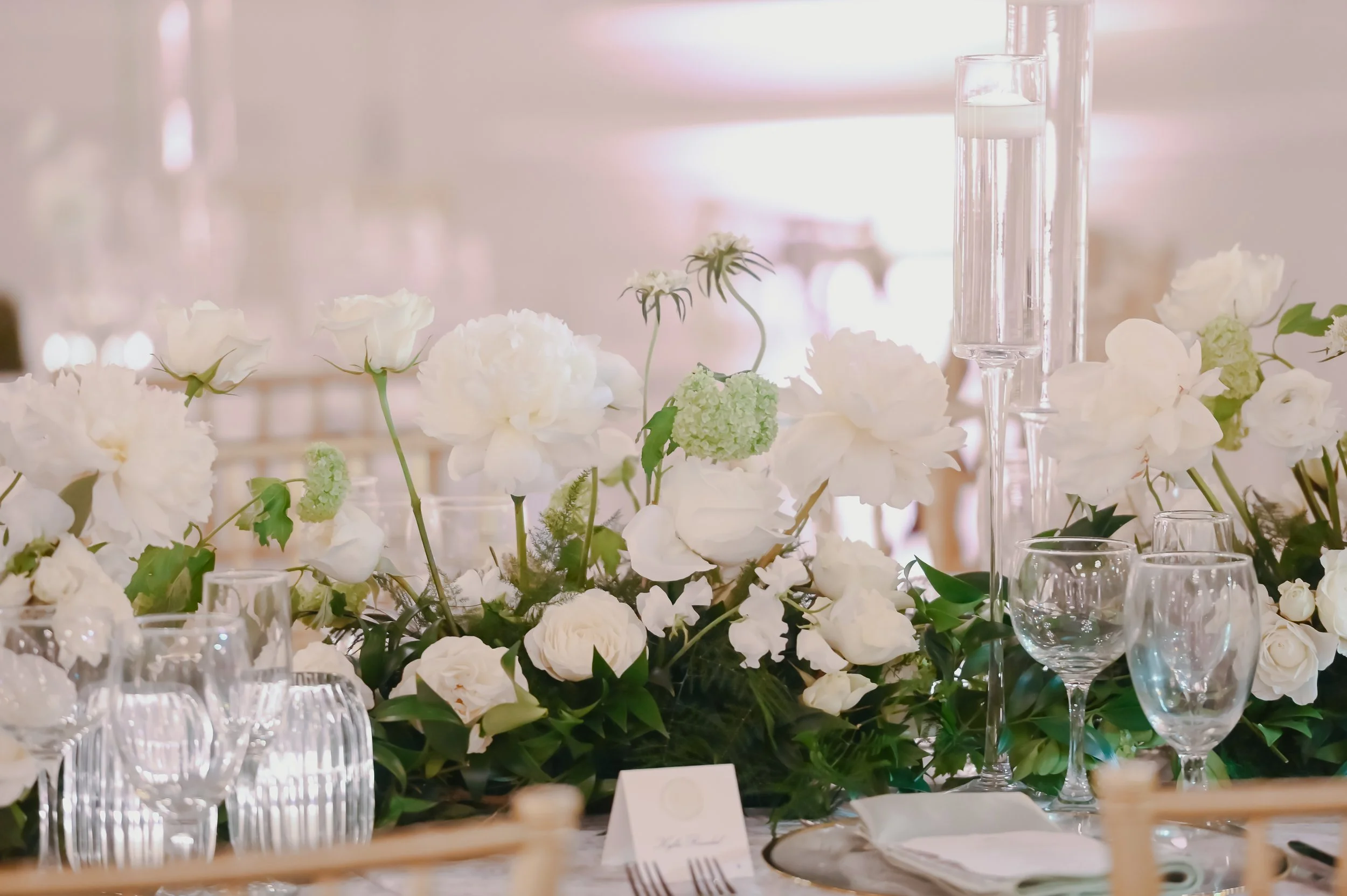 A floral centerpiece consisting of white and green flowers on a table set for a formal event with glassware and utensils.