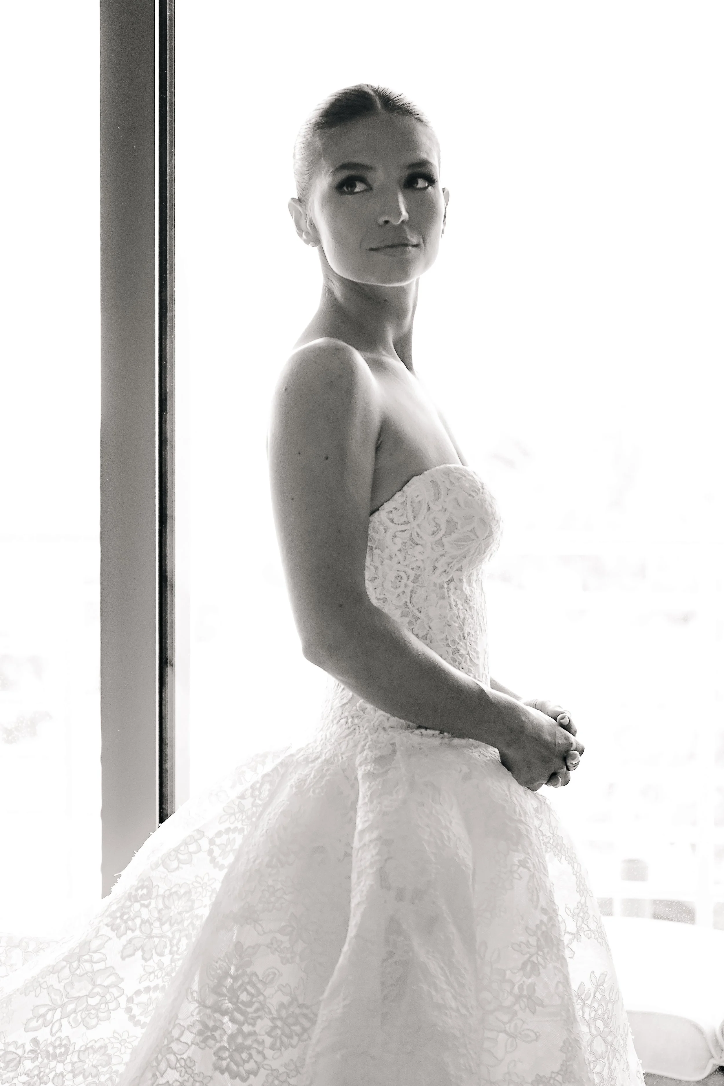 A woman in a wedding dress sitting by a window, looking to her left, with her hands clasped in her lap, in black and white photograph.