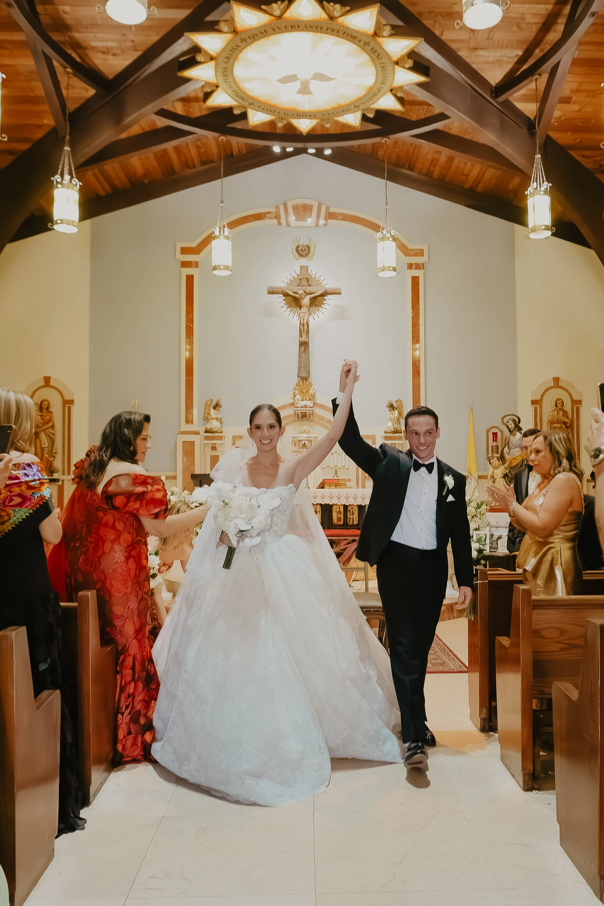 A newly married couple exiting a church, holding hands with their arms raised in celebration. The bride wears a white wedding gown and holds a bouquet of white flowers; the groom wears a black tuxedo. Guests are clapping and smiling as they walk down