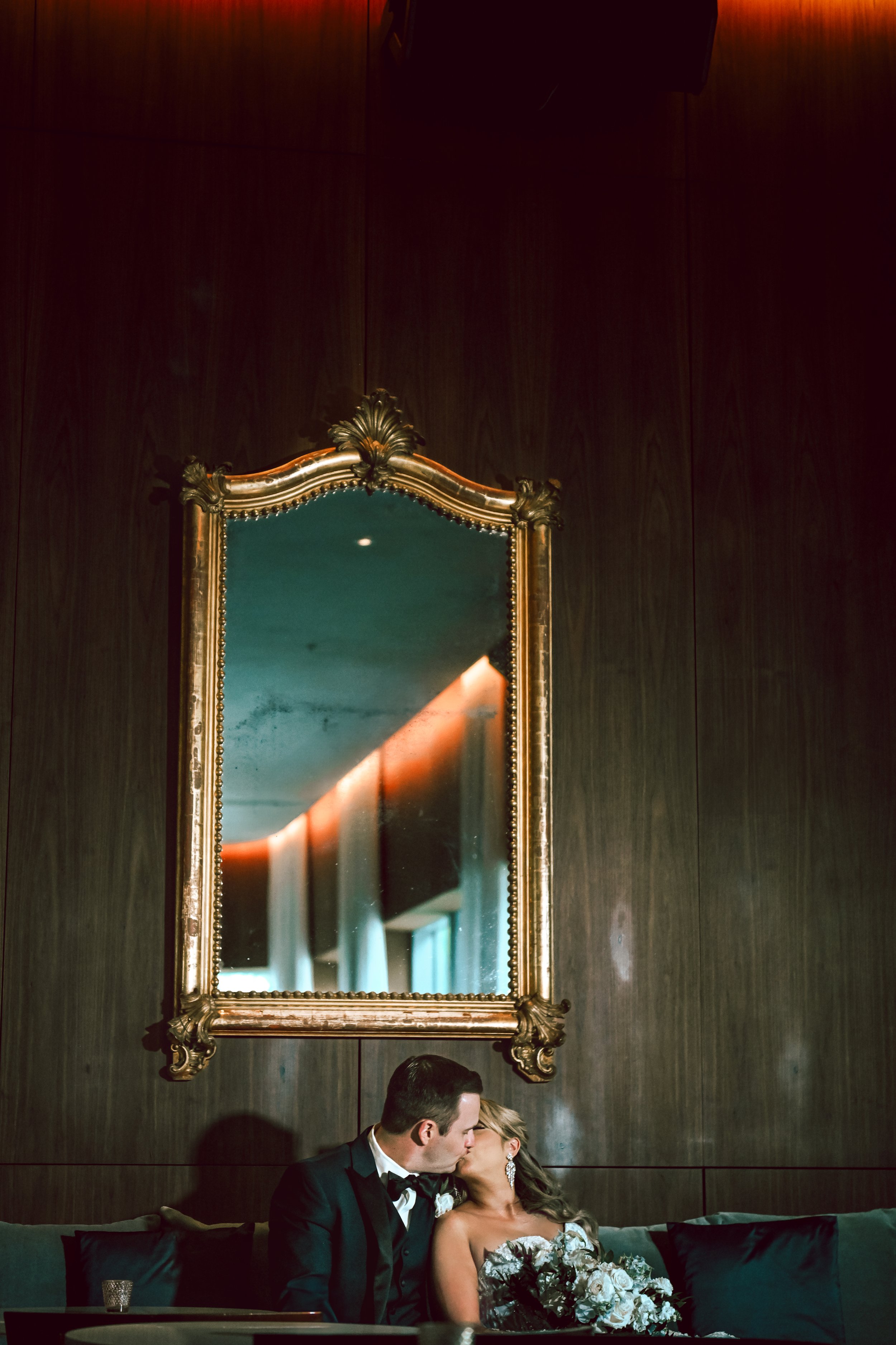 A bride and groom sit on a dark green couch, kissing, in an elegant room with dark wood paneling and a large ornate mirror hanging above them. The bride holds a bouquet of white and green flowers, and both are dressed formally, with the groom in a tu