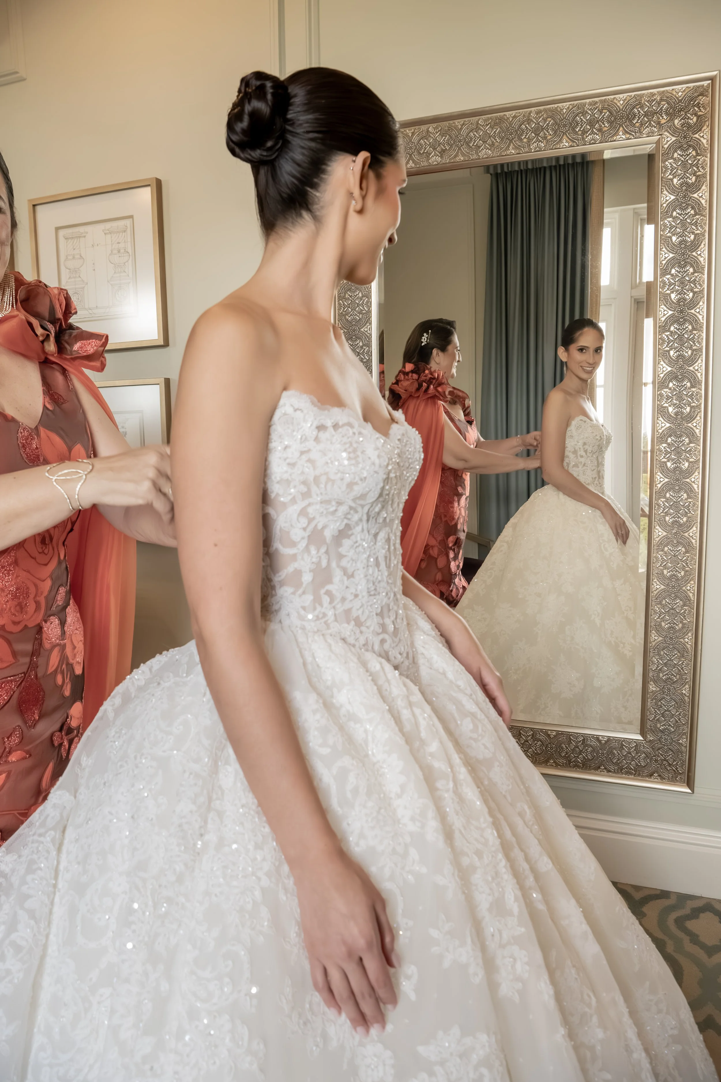 A bride getting ready in front of a mirror with an elegant gold frame. She is wearing a white wedding gown with intricate lace details.