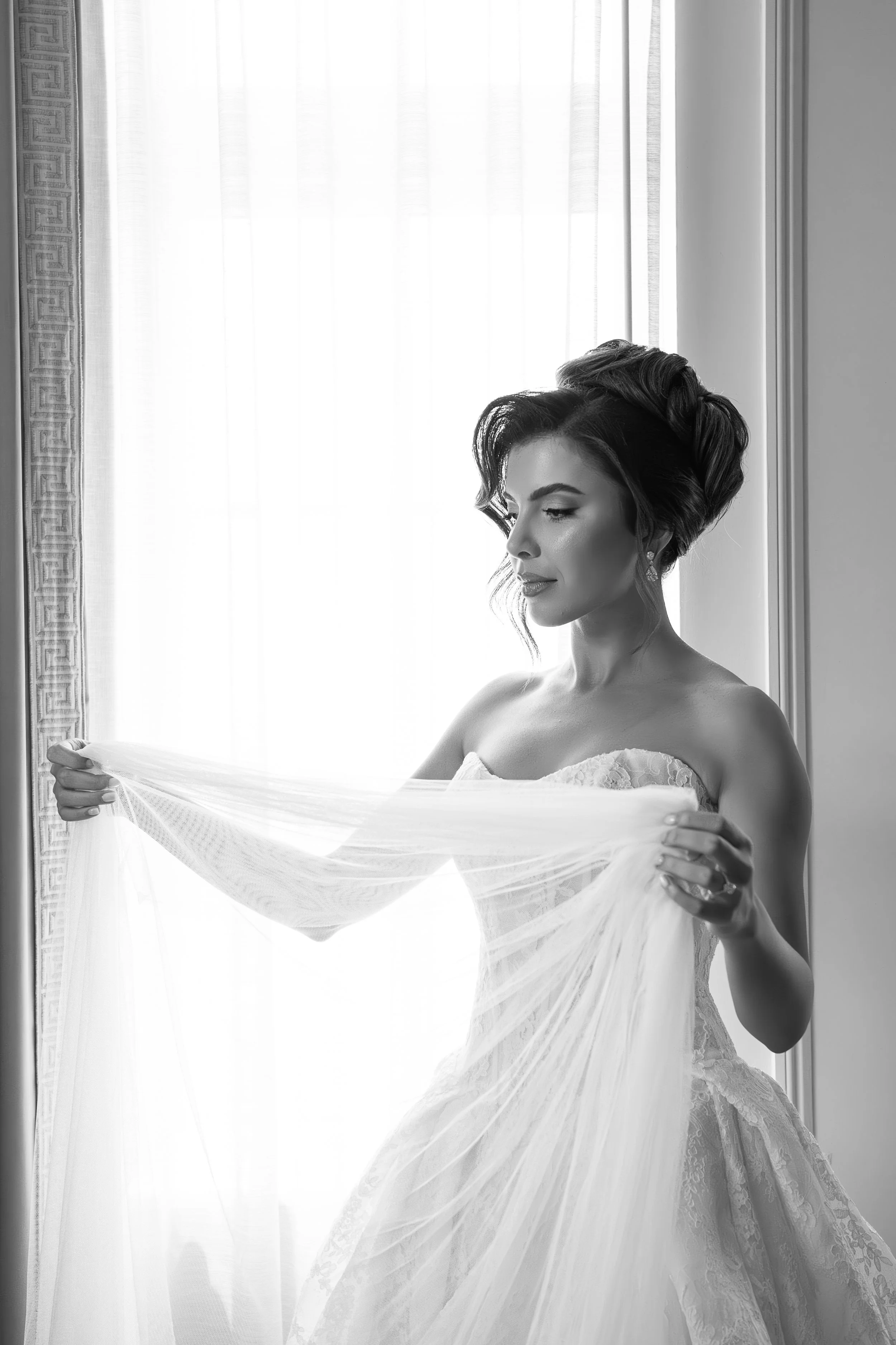 A woman in a wedding dress stands by a window, holding a sheer fabric, with sunlight streaming in.