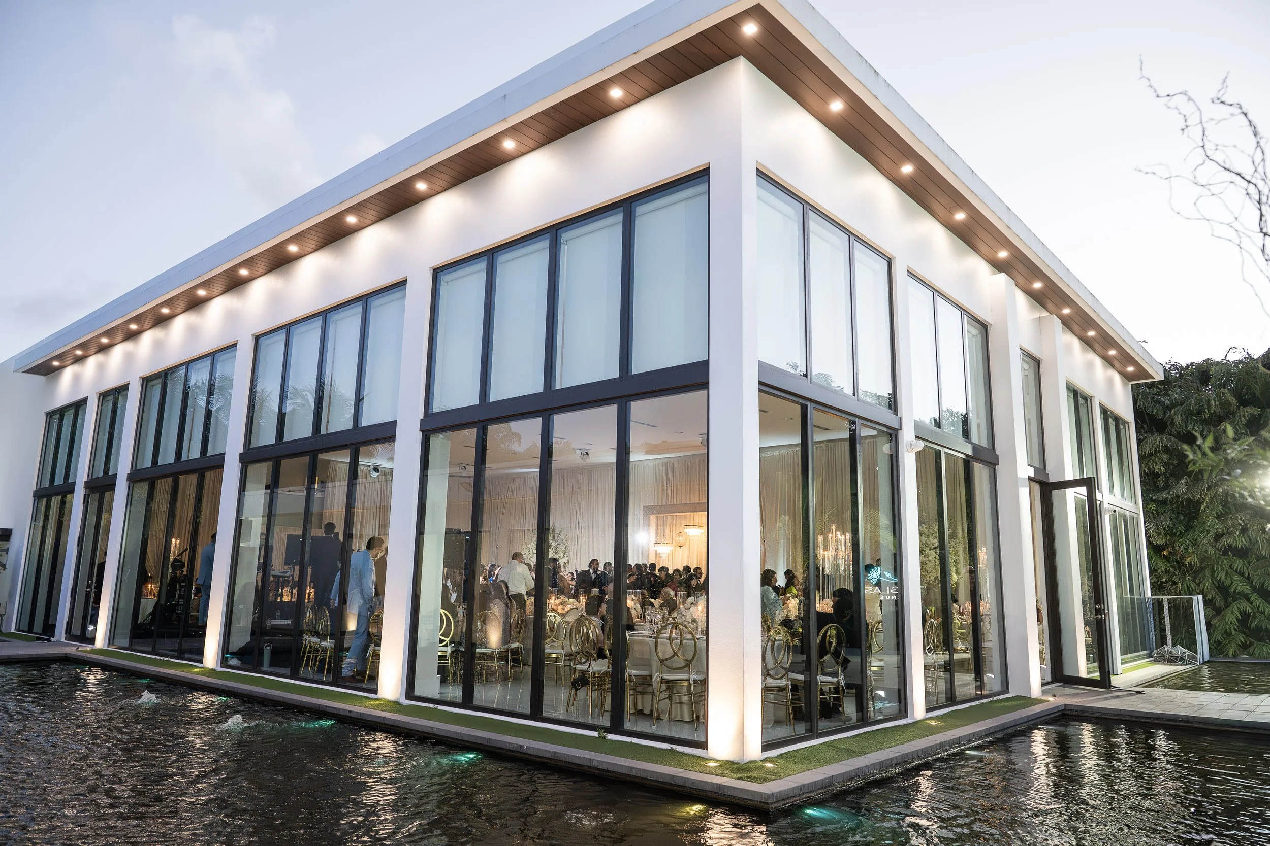 Modern white building with large glass windows and a reflecting pool surrounding it, during evening with the lights inside on, and people gathered inside for a social event.