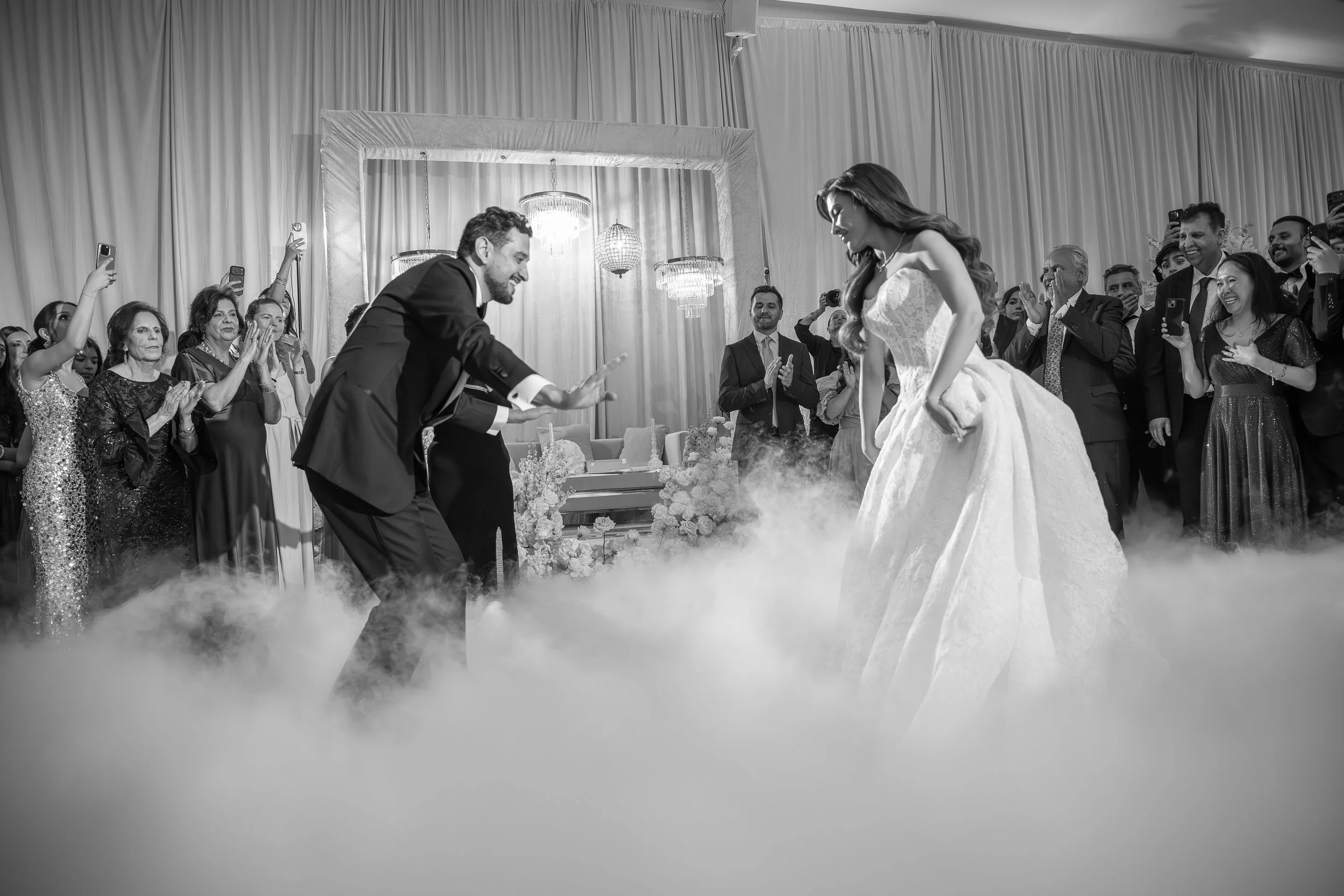 A black and white photo of a wedding scene with a couple in the center, the groom in a tuxedo and the bride in a wedding gown, surrounded by guests. The groom and bride are smiling and appear to be enjoying a dance or moment together. Guests are clap