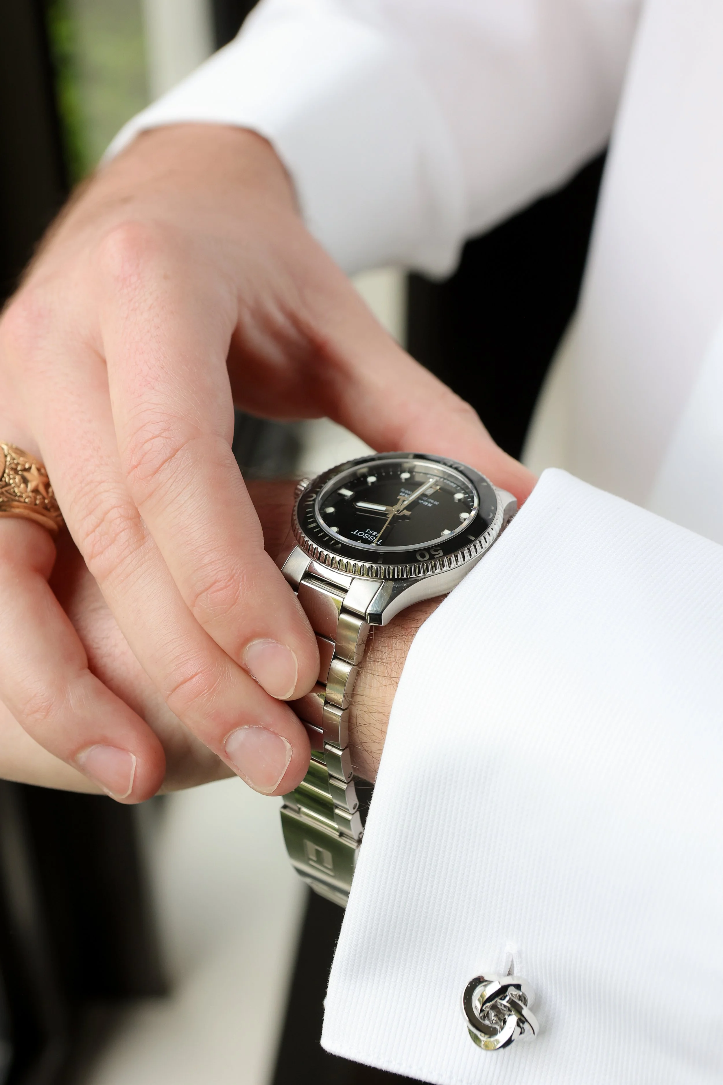 Close-up of a person's wrist wearing a silver wristwatch, with their hand adjusting the watch. The person is dressed in a white shirt with a French cuff and a cufflink featuring interlocked G's. The background is blurred.