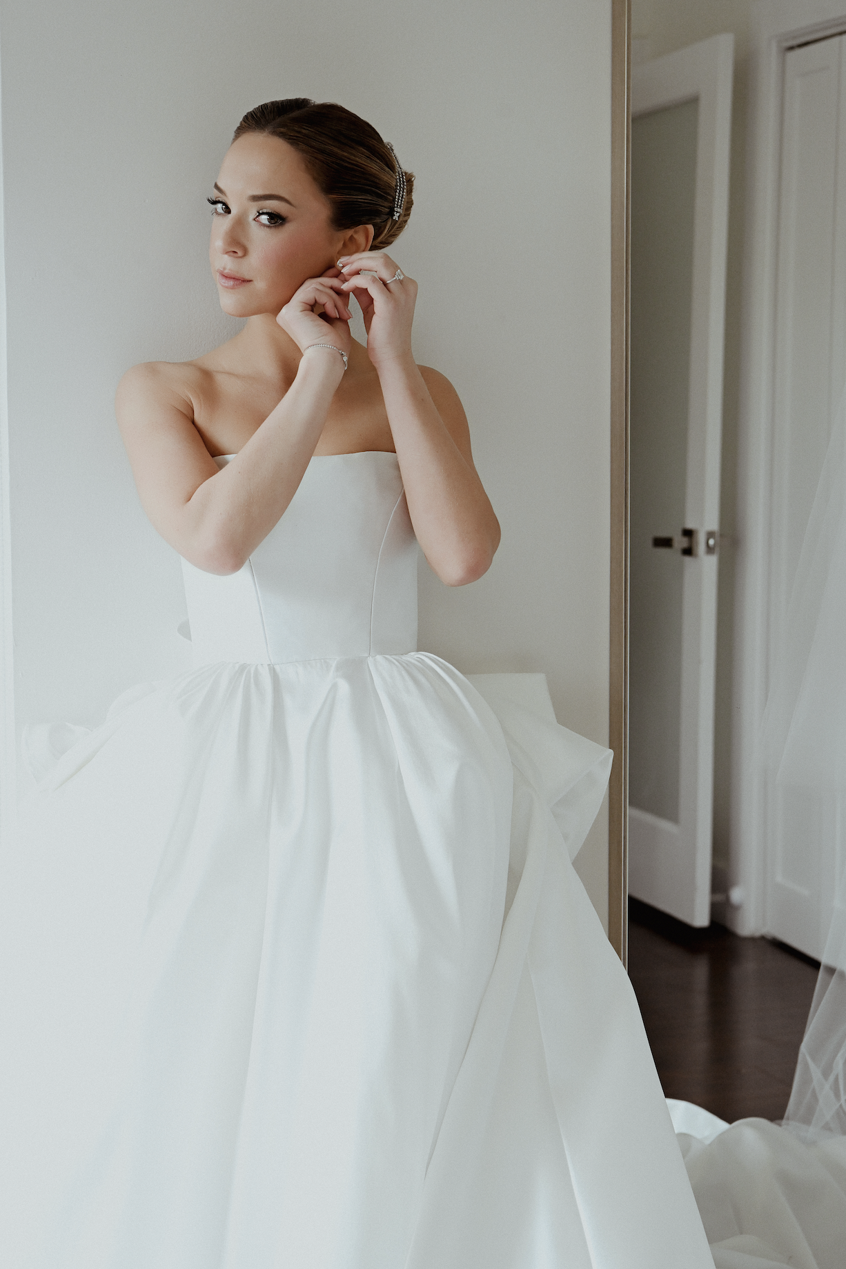 Brunette woman in a wedding dress adjusts her earring in front of a mirror.