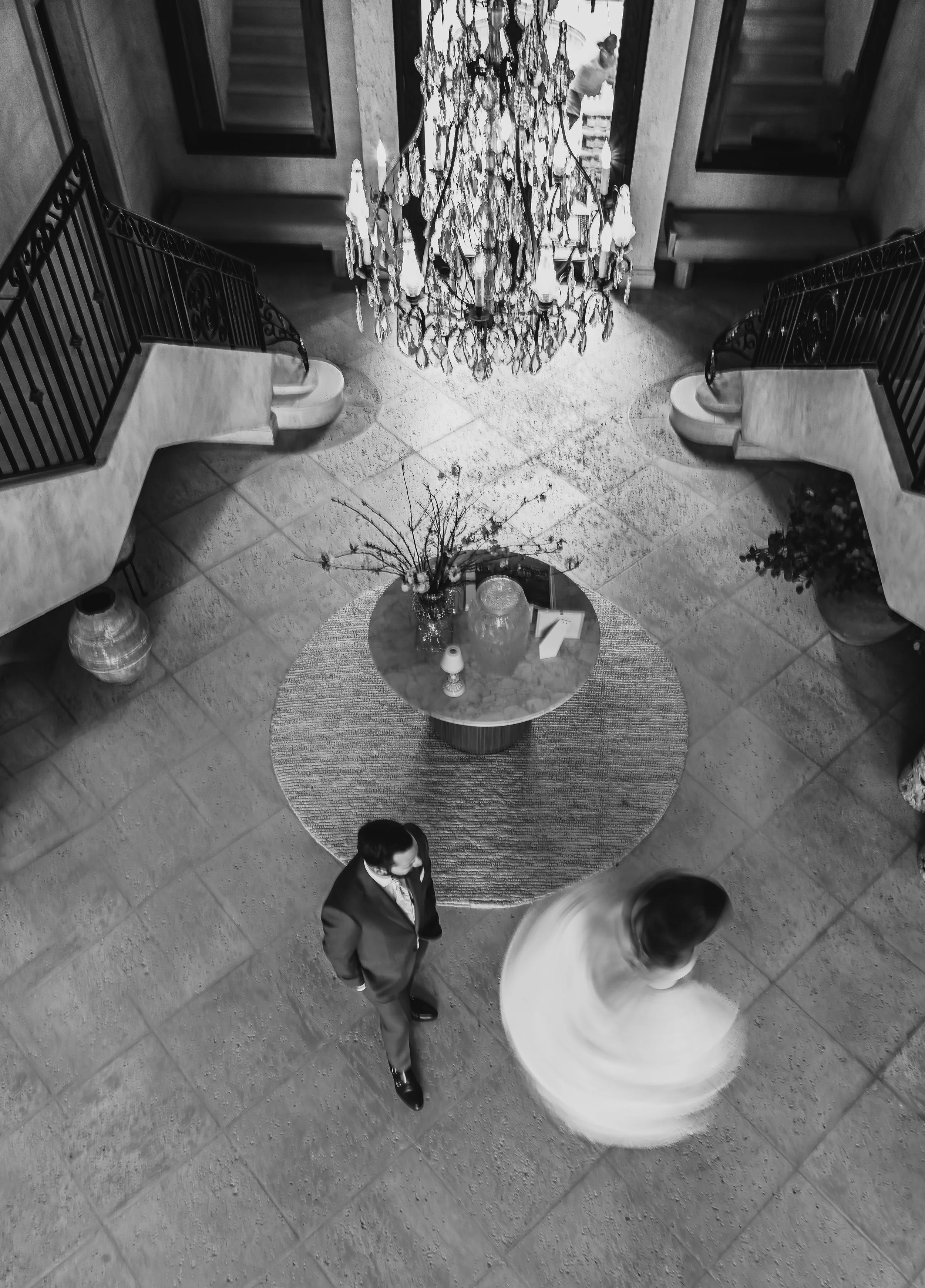 A grand chandelier hangs from the ceiling in a luxurious lobby, with two people dressed in formal attire walking underneath. The space includes stairs, mirrors, and decorative plants.