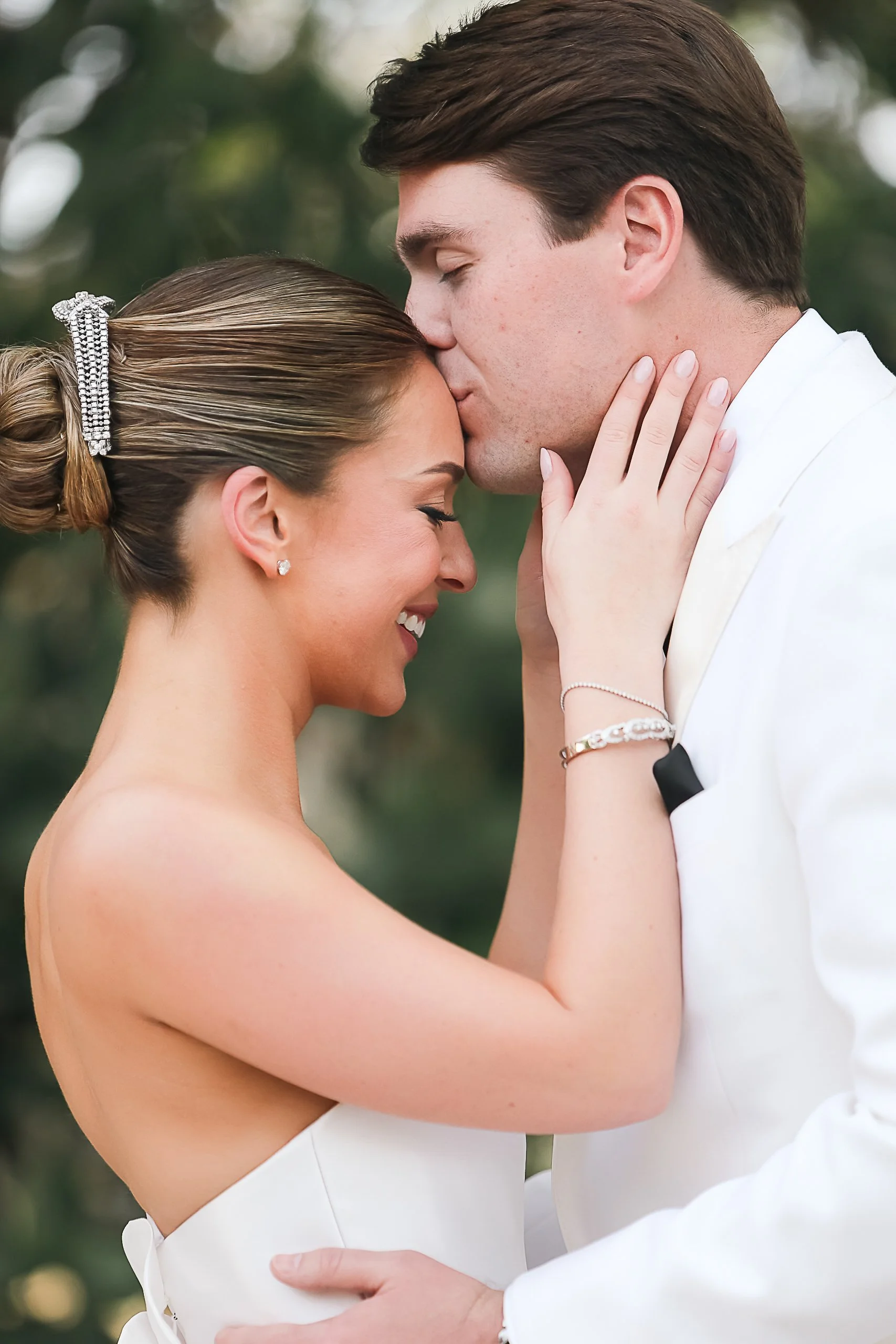 A couple dressed in wedding attire sharing a kiss outdoors, with the groom gently holding the bride's face and the bride smiling with closed eyes.